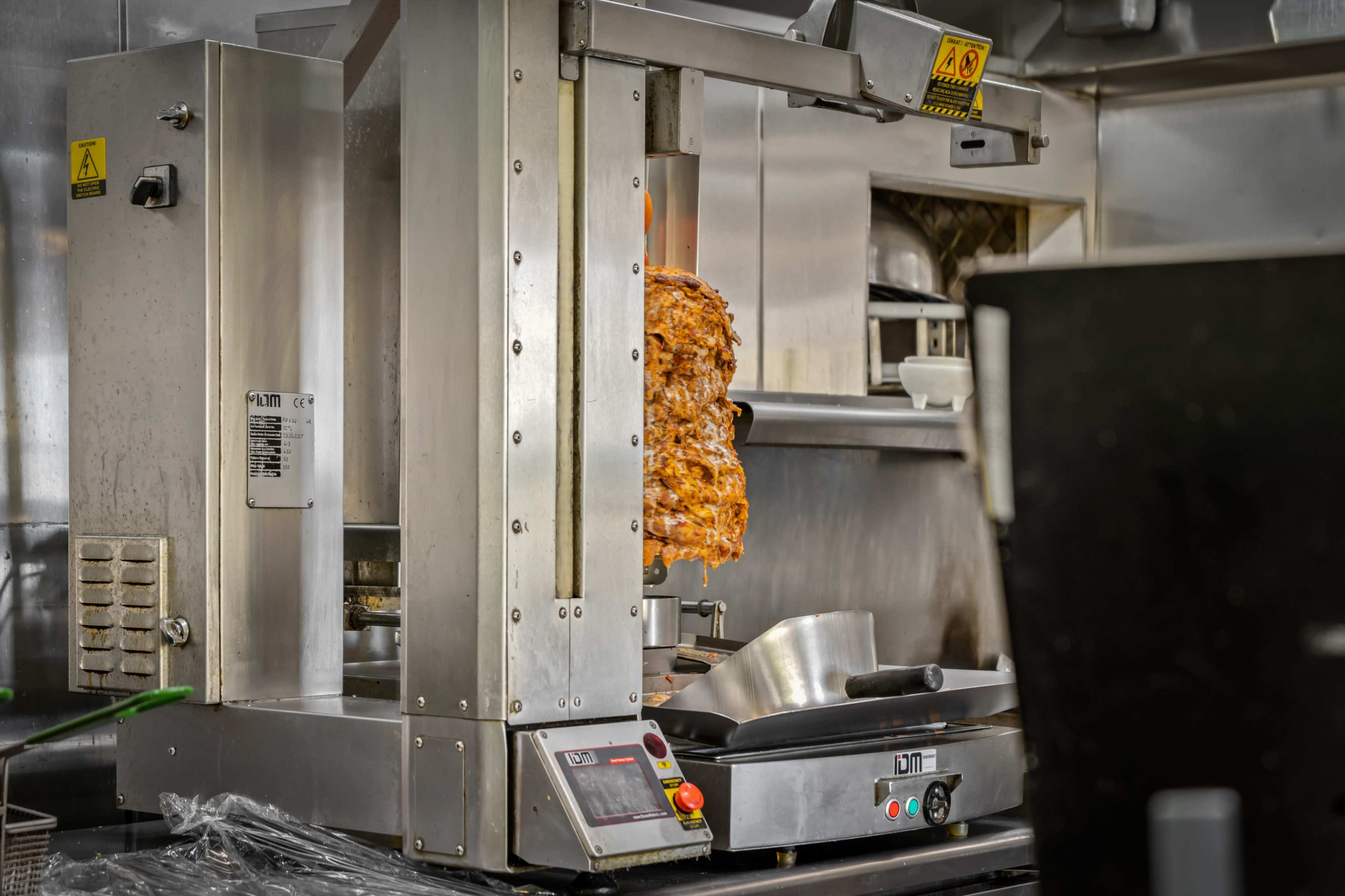 A vertical rotisserie displays cooked meat in a commercial kitchen, with metallic surfaces and cooking equipment visible in the background.