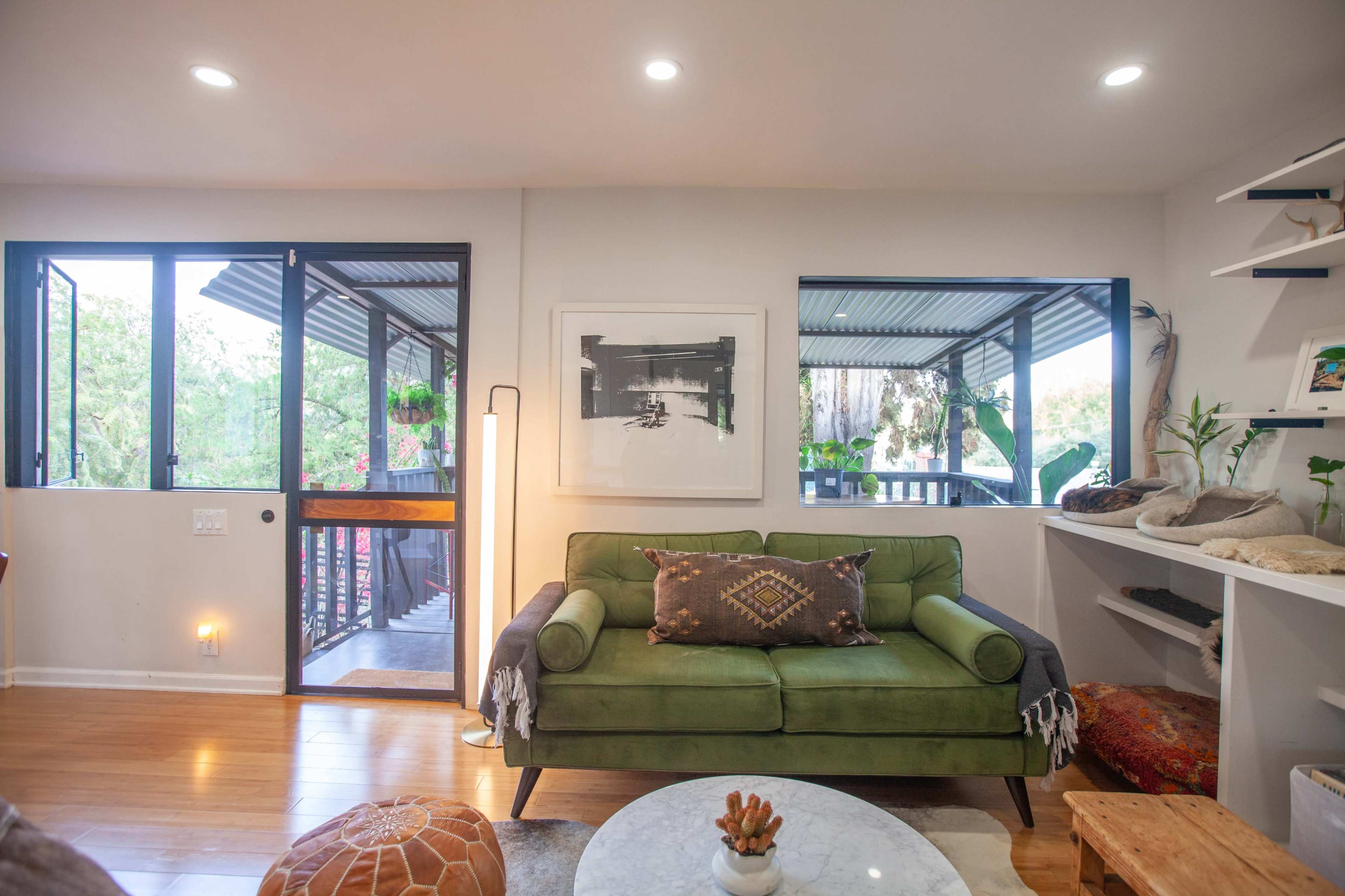 A cozy living room featuring a green sofa, a round coffee table, and large windows allowing natural light, with a view of an outdoor balcony.