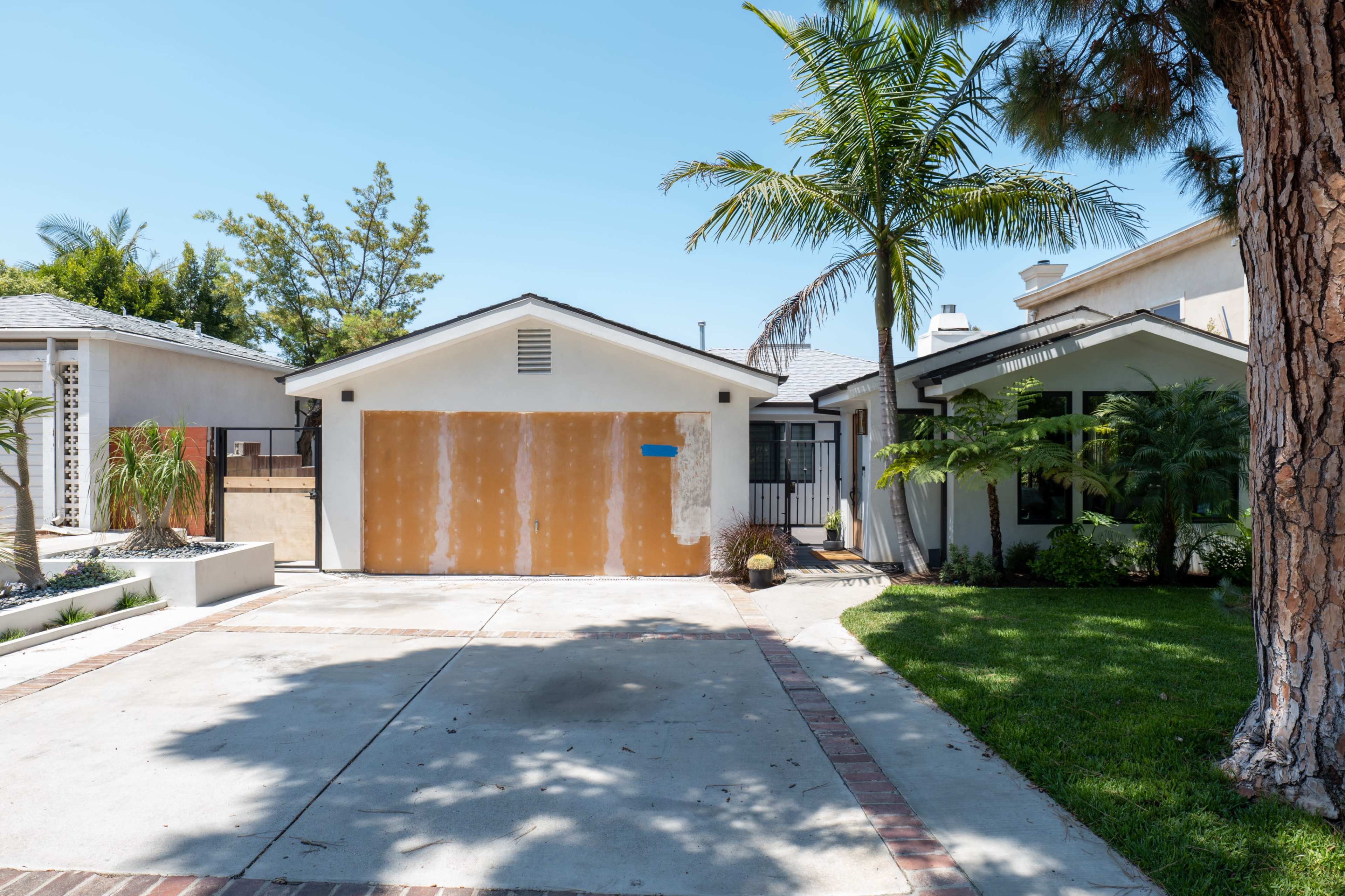 A residential property with a driveway leading to a garage with an incomplete paint job and palm trees in the front yard.