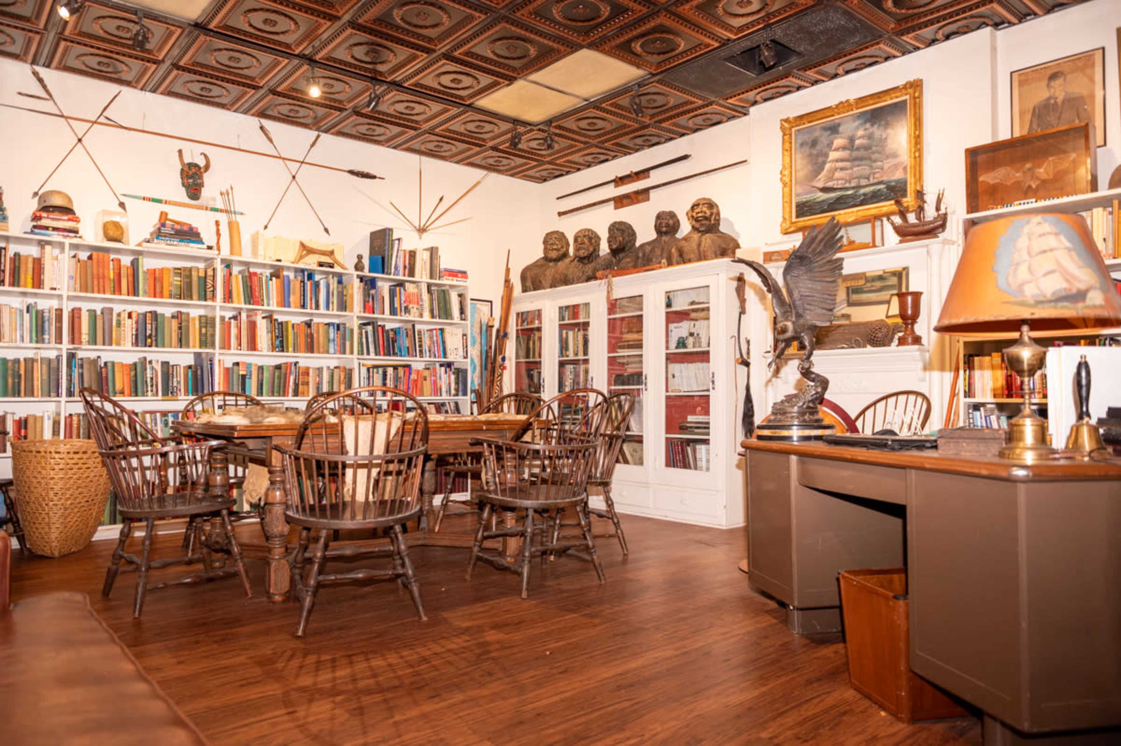A well-furnished room with a wooden table surrounded by chairs, a large bookshelf filled with books, and various artworks and sculptures displayed on the walls and shelves.