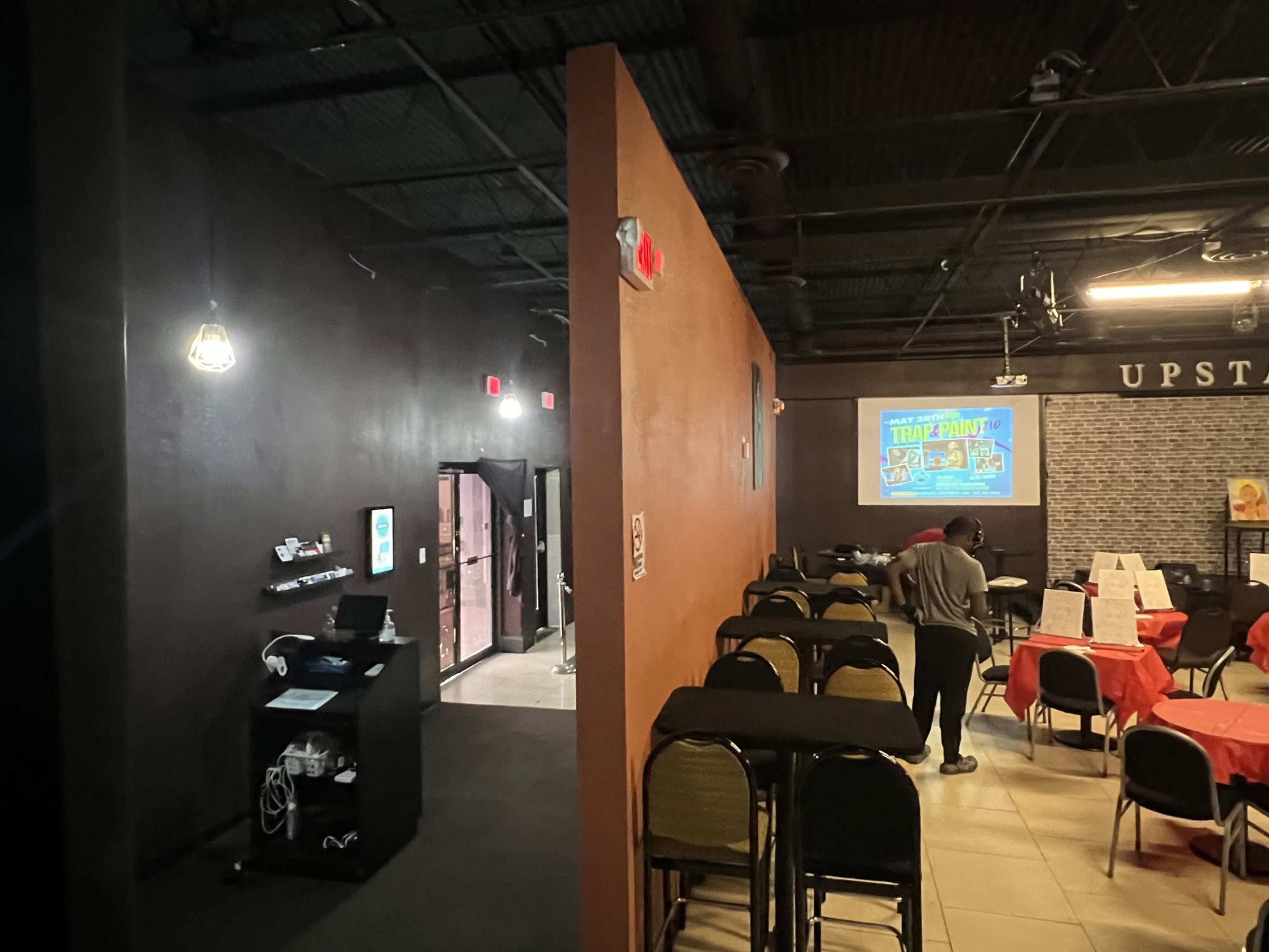 A dimly lit room with a dark wall on one side, tables and chairs arranged for dining, and a screen displaying an event poster in the background.