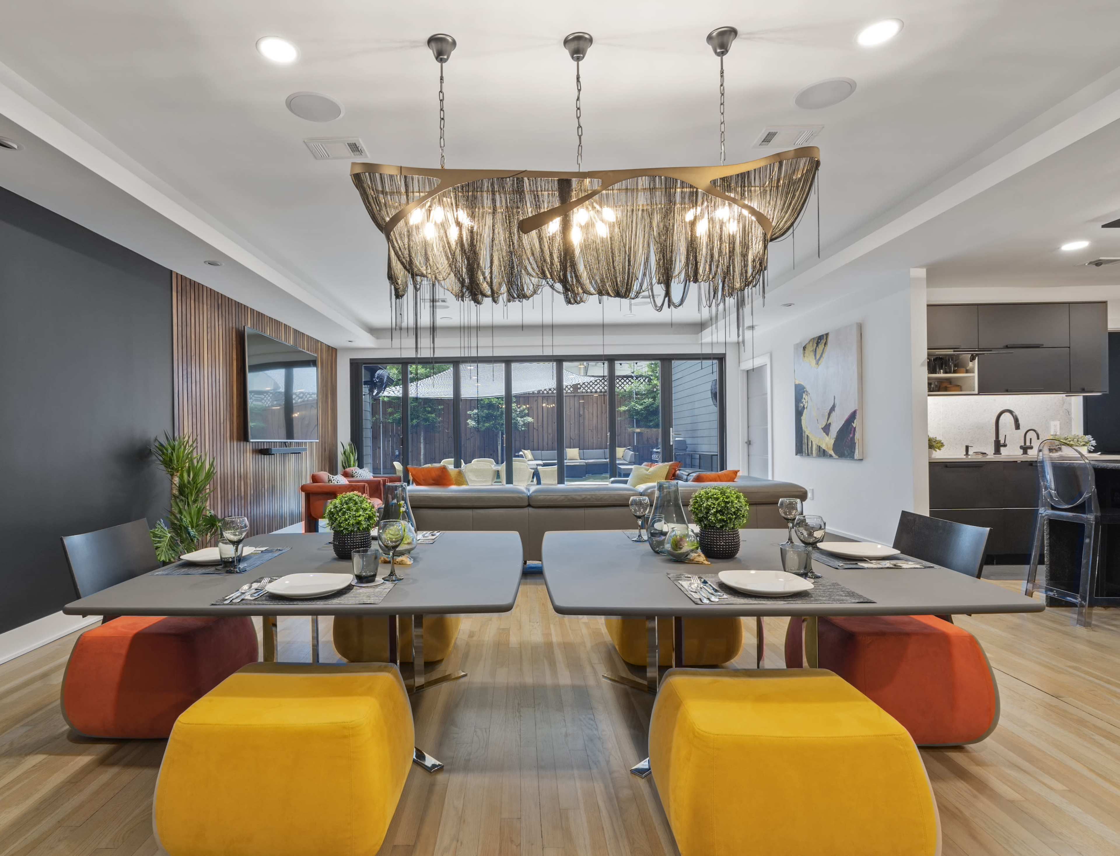 A contemporary dining area features two modern tables with colorful seating, a unique light fixture, and large windows overlooking an outdoor space.