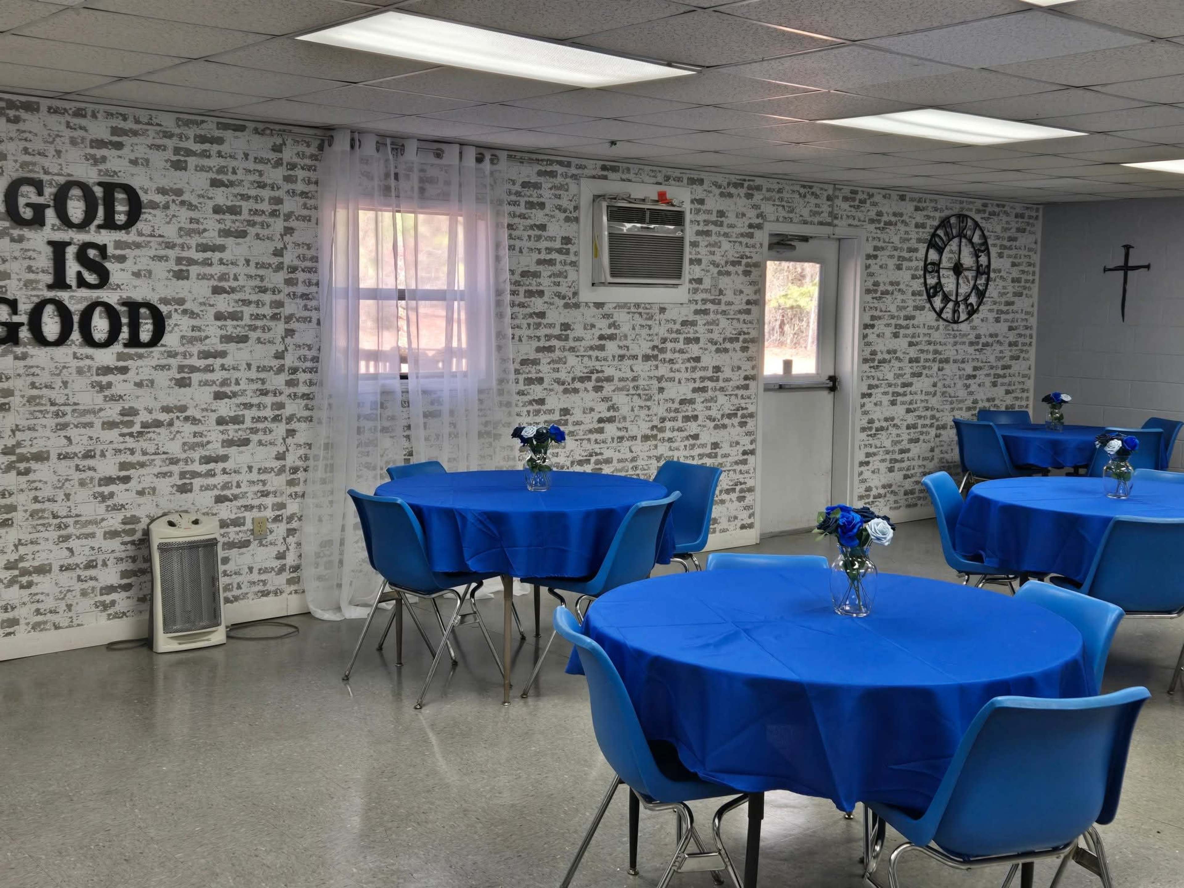 The image shows a room with blue tablecloths on round tables, decorated with small flower vases, and a wall featuring a pattern of text that includes the phrase "GOD IS GOOD."