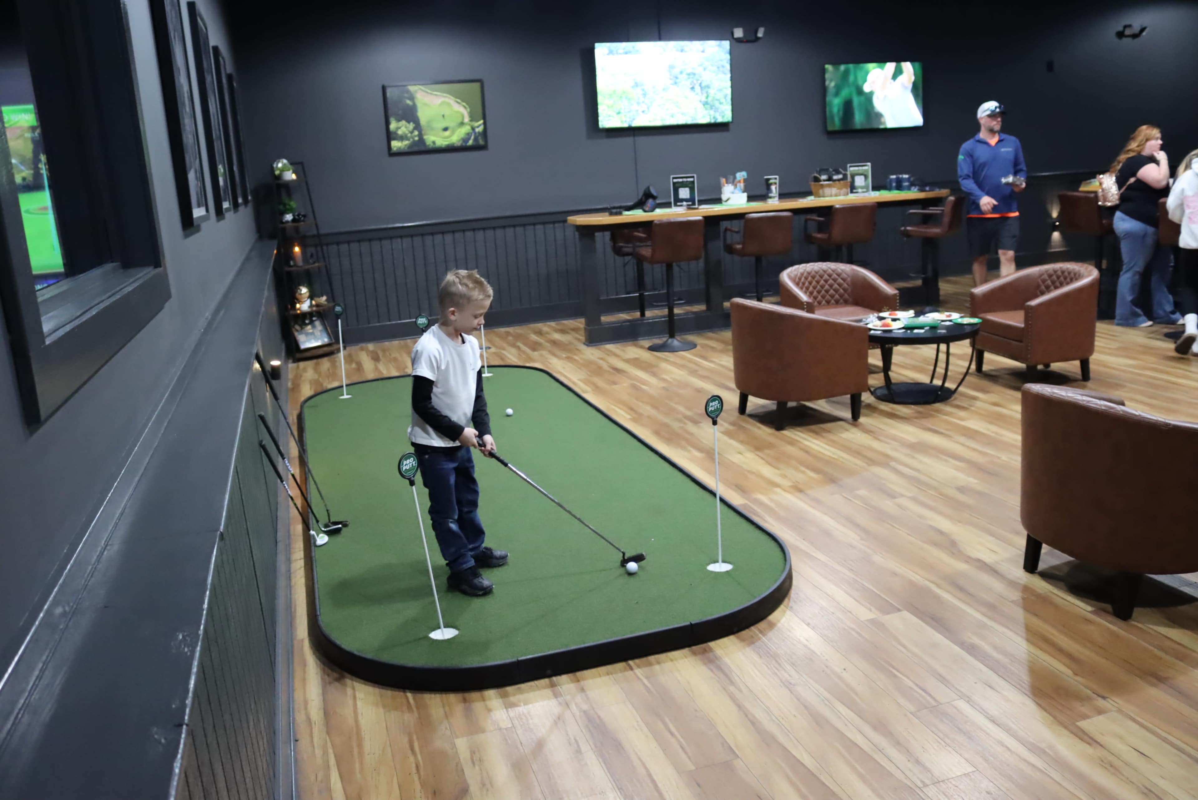 A child stands on a putting green indoors, preparing to take a shot while several people socialize at tables in the background.