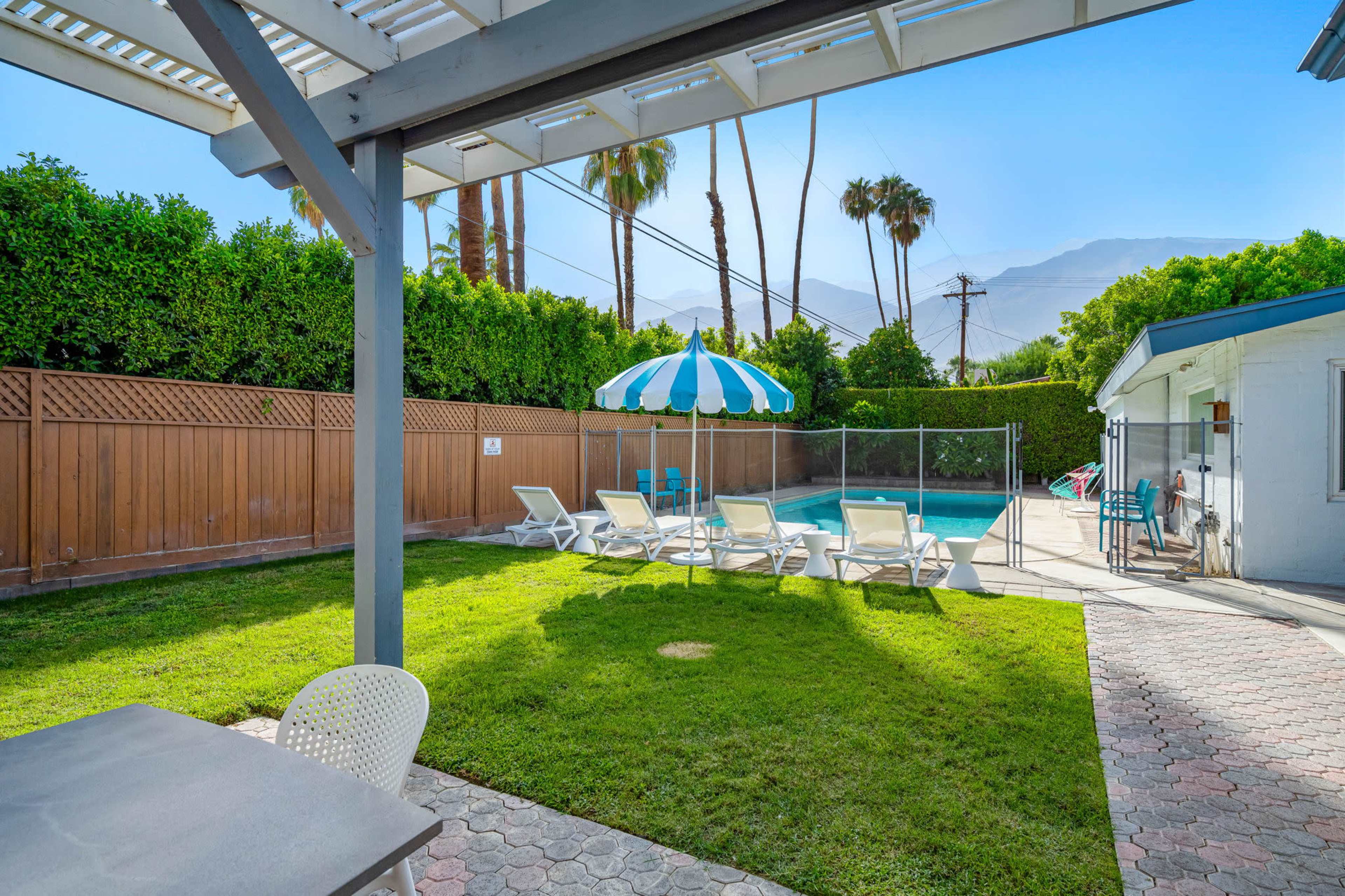 A backyard features a swimming pool surrounded by lounge chairs and a large umbrella, with palm trees and a wooden fence in the background.
