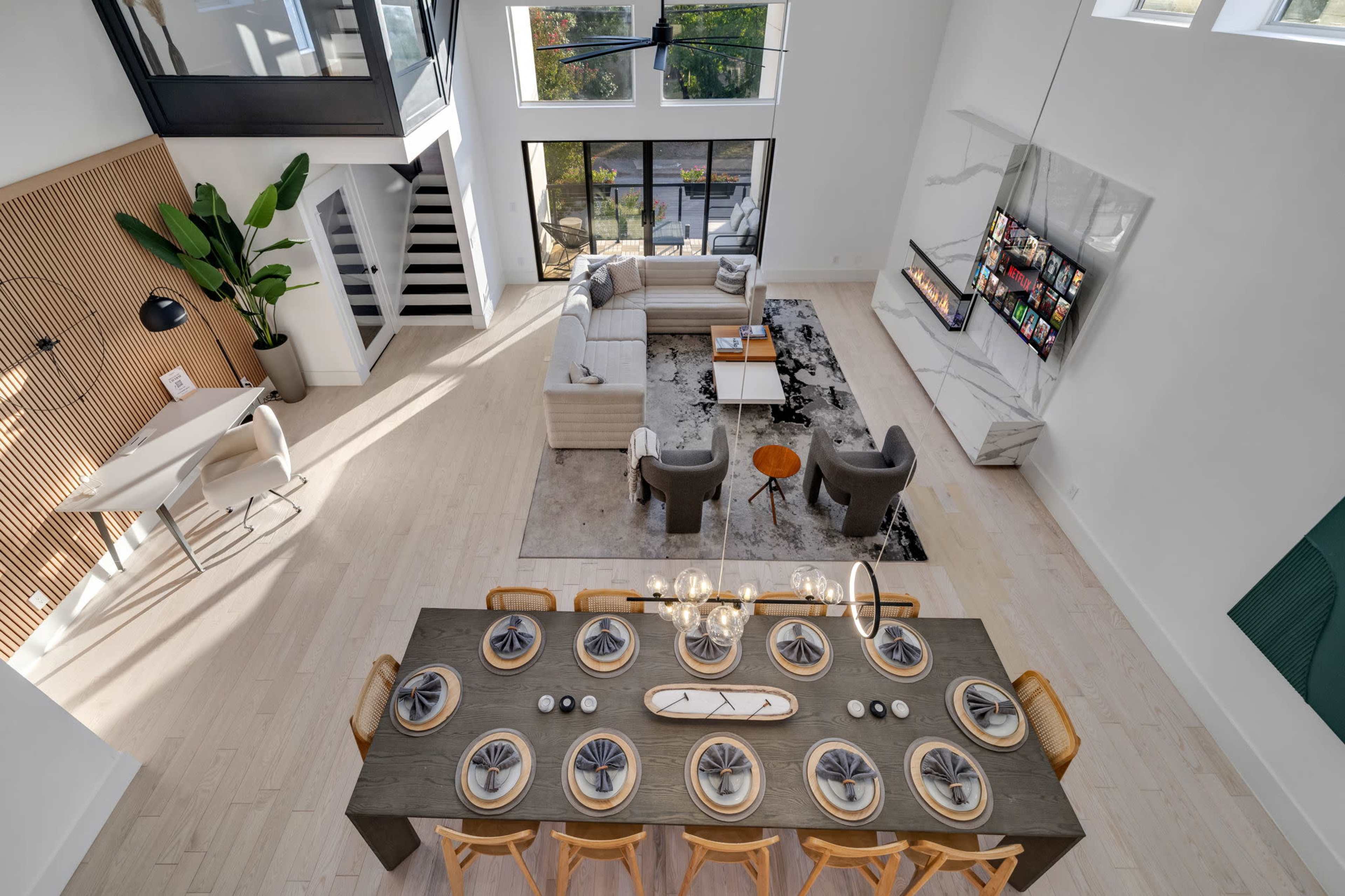 The image shows a modern, spacious living and dining area with a large table set for a meal, stylish furniture, and large windows allowing natural light to fill the room.