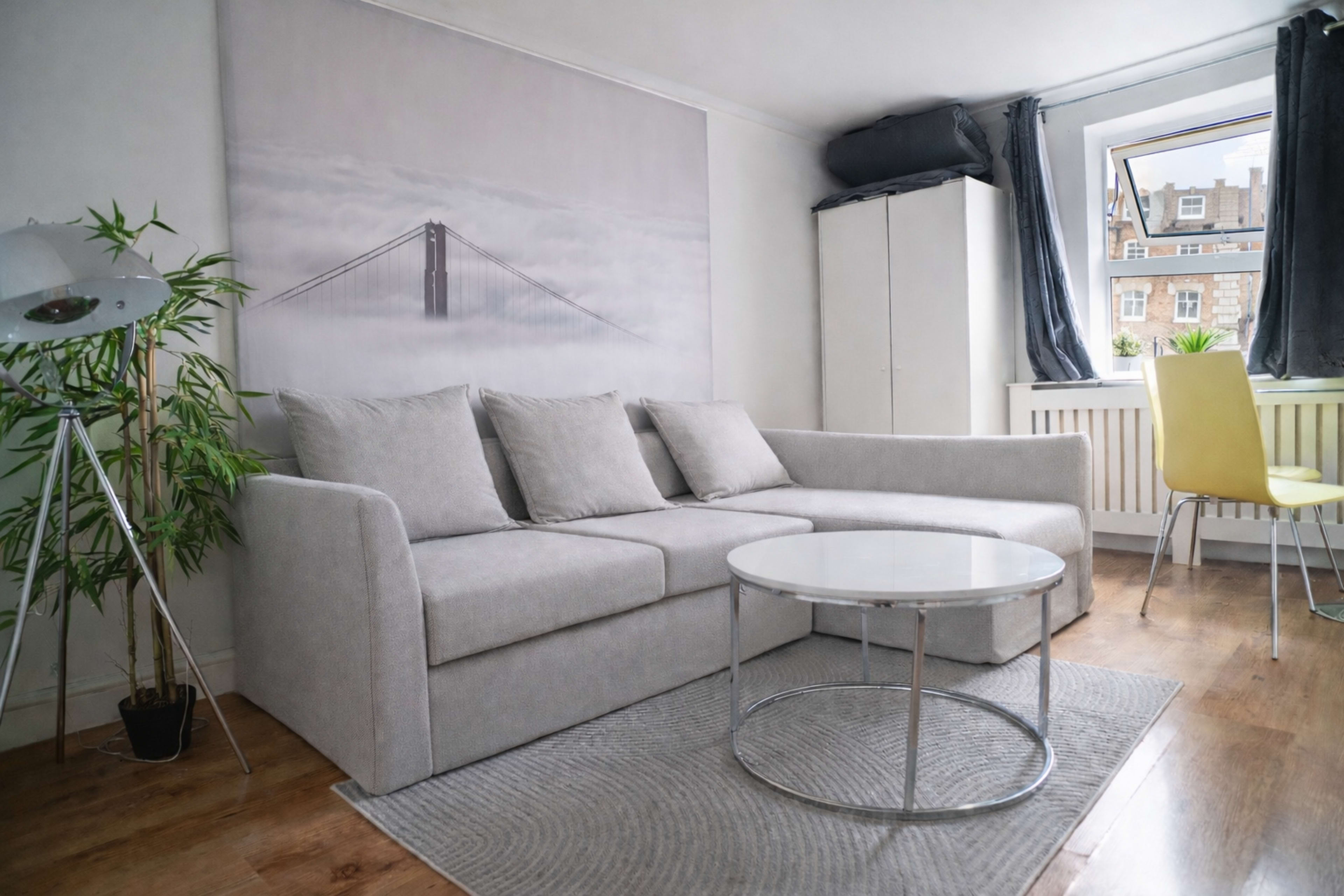 The image shows a modern living room with a gray sectional sofa, a round glass coffee table, and a large wall mural of a bridge.