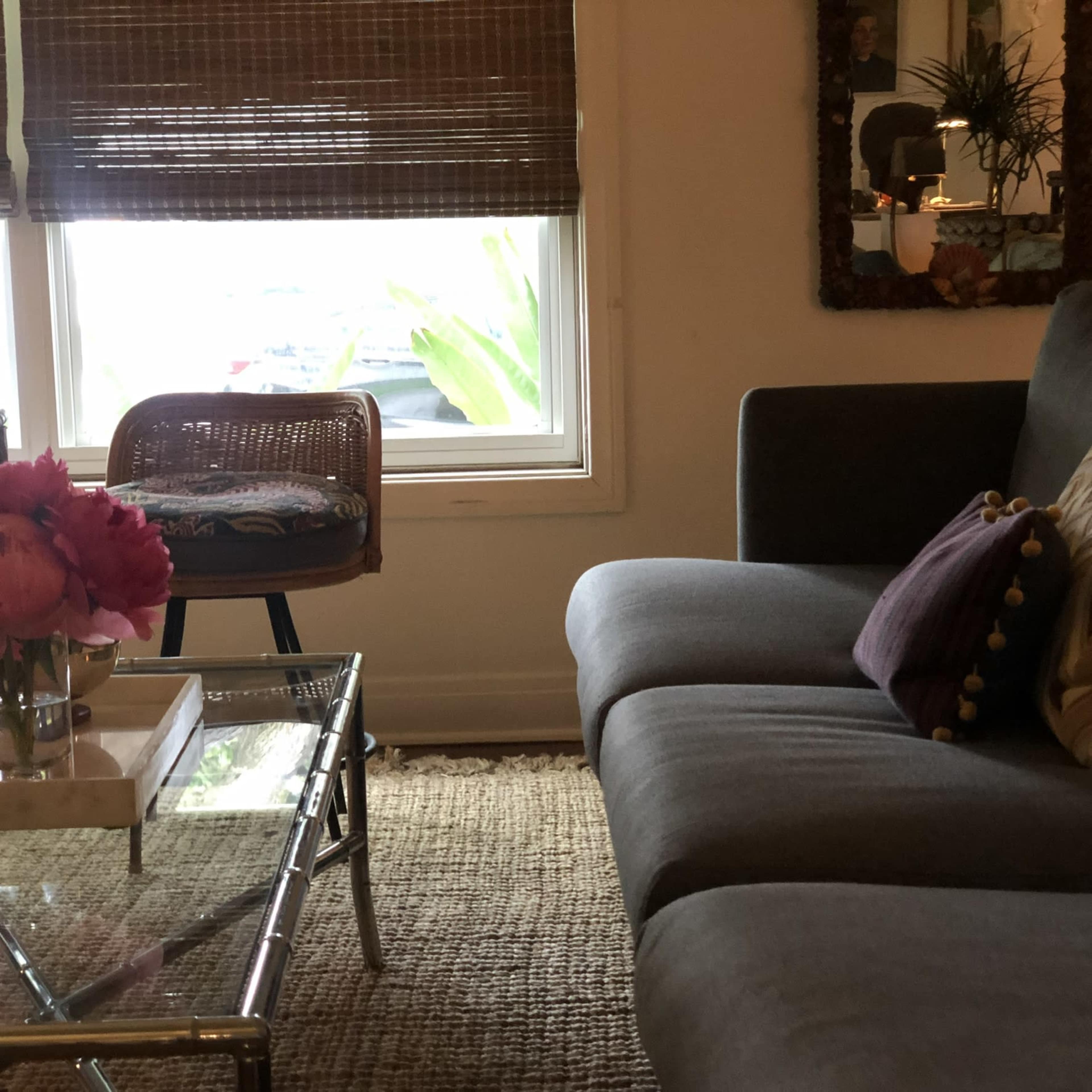 A gray sofa and a glass coffee table are positioned in a cozy room with a large window displaying greenery outside.