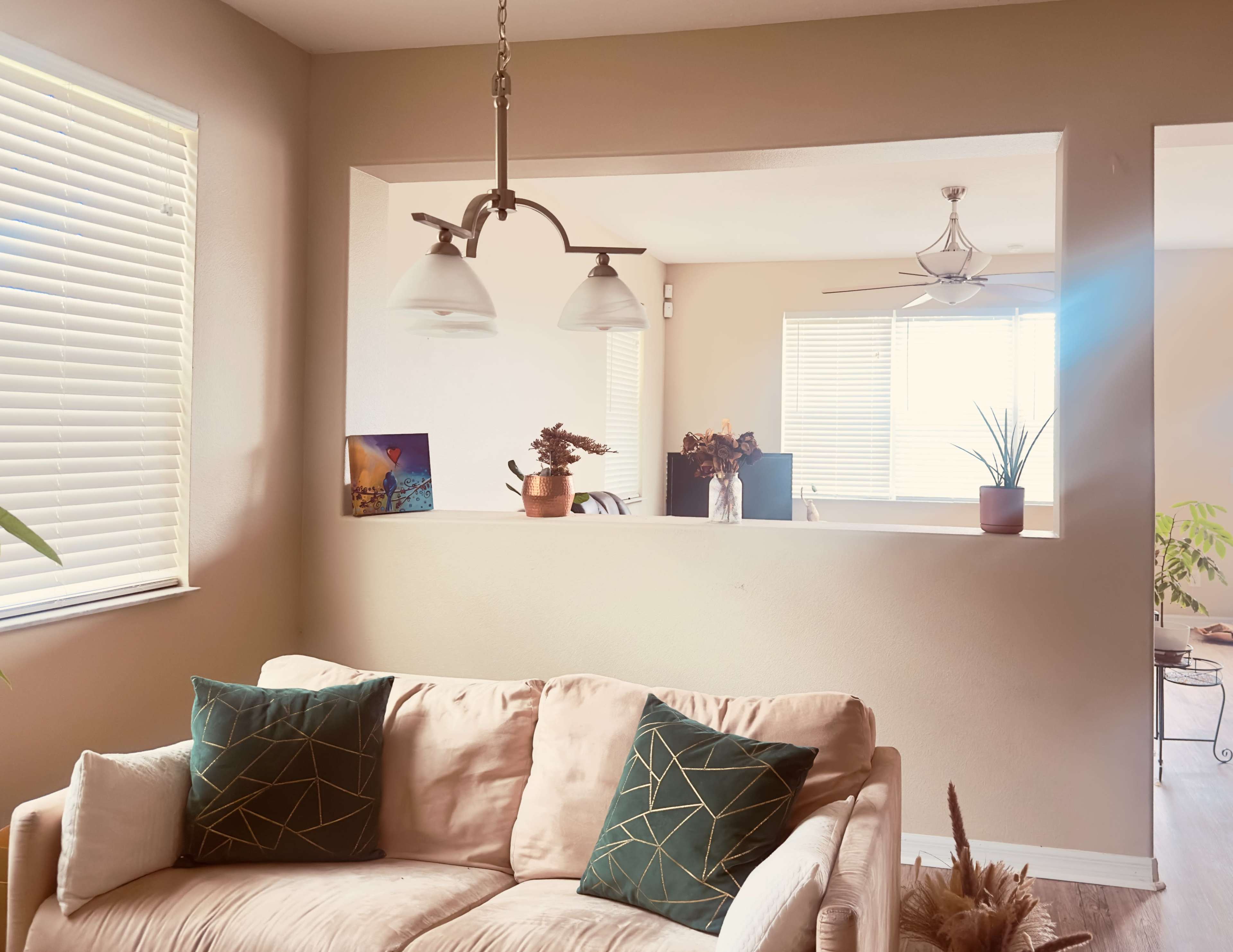 The image shows a living area with a beige couch adorned with green geometric-patterned pillows, a pendant light hanging above, and a view of a bright room featuring plants and natural light filtering through windows.
