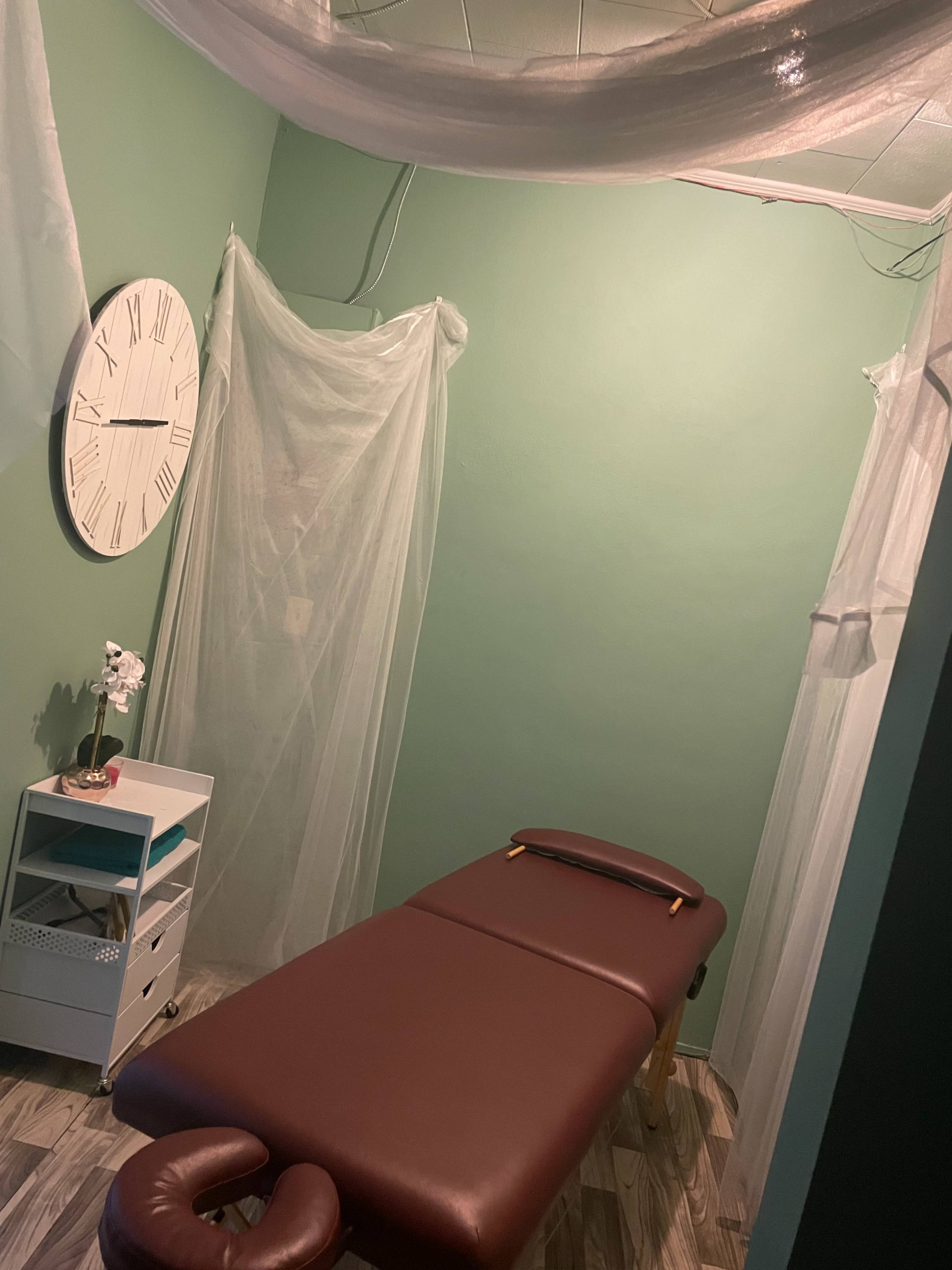 A massage room with a brown massage table, a wall clock, and sheer curtains hanging in a green-walled space.