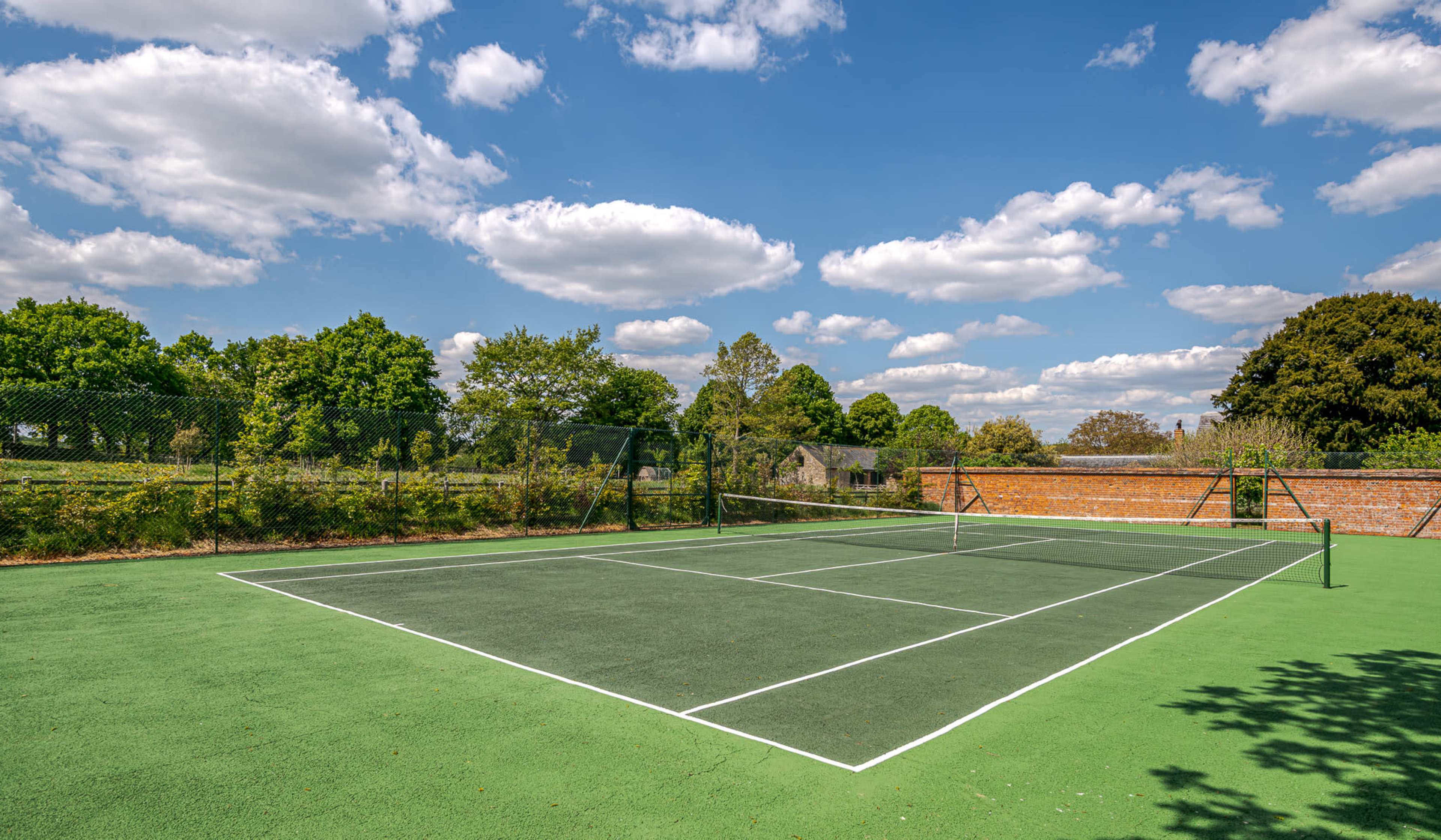 A tennis court with green playing surface is surrounded by trees and under a blue sky with scattered clouds.