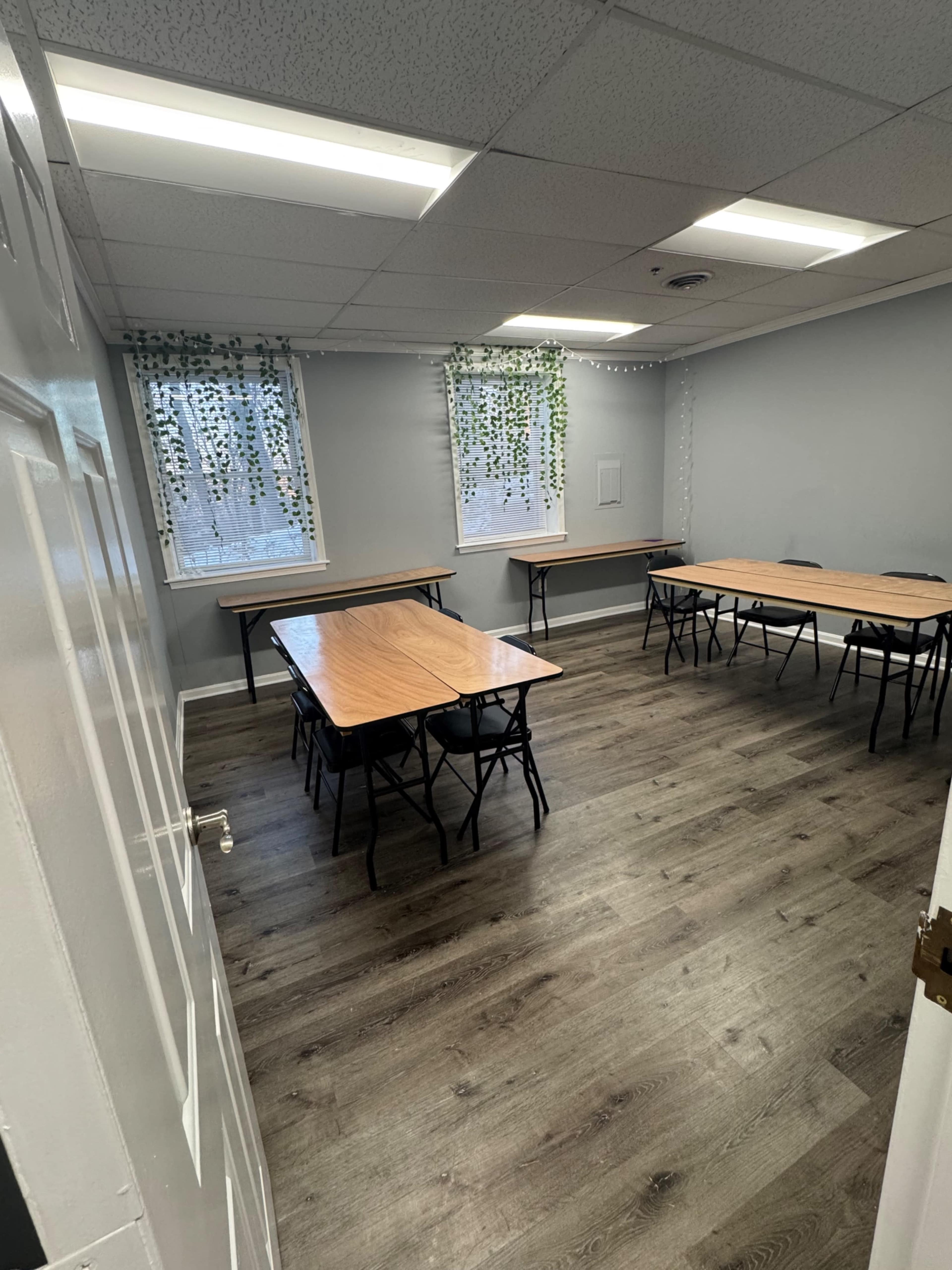 The image shows a classroom featuring several wooden tables arranged around the room, with green plants hanging in front of two windows.