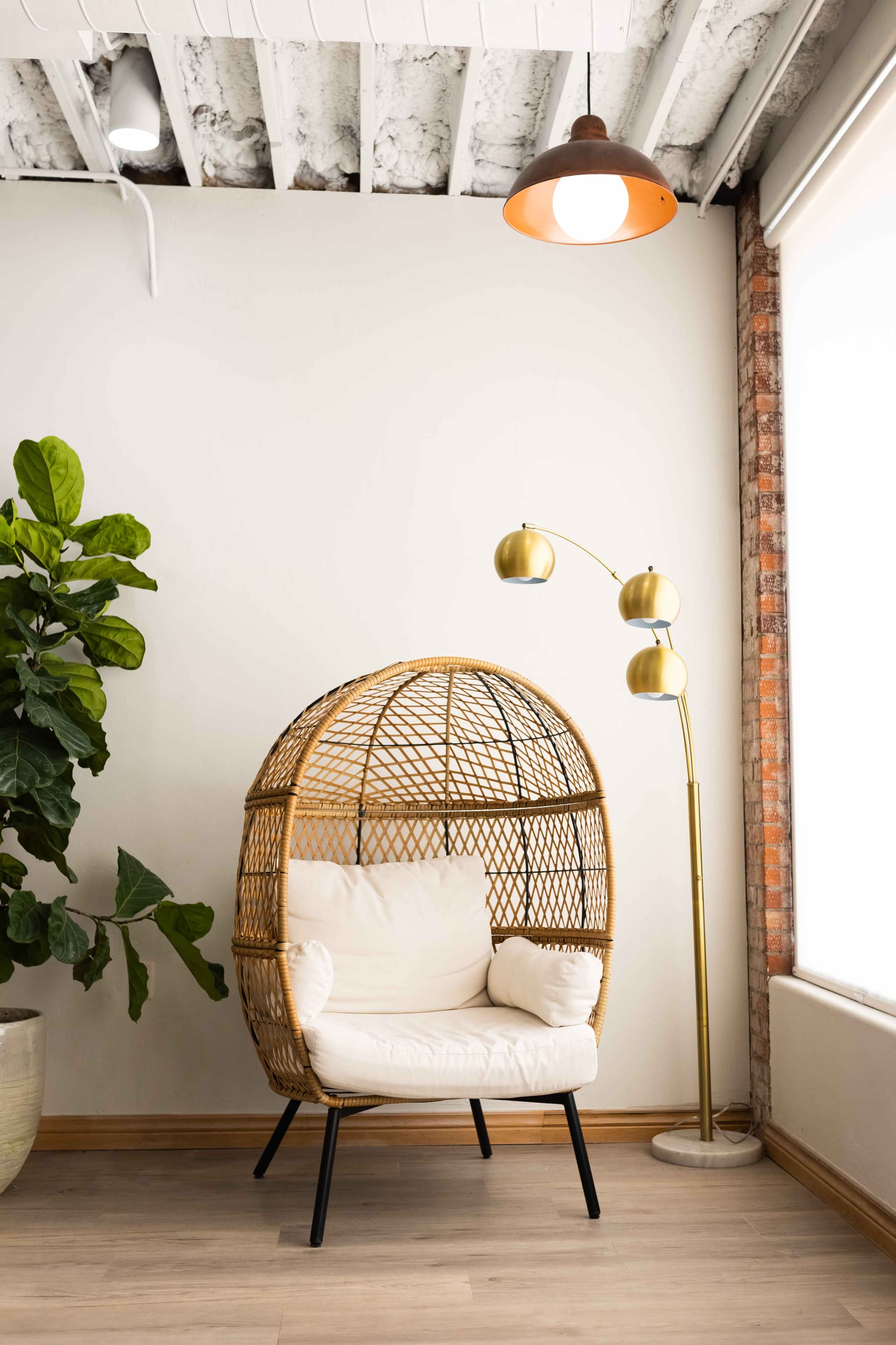A wicker chair with a soft cushion is situated next to a modern floor lamp in a room with exposed brick and a potted plant.