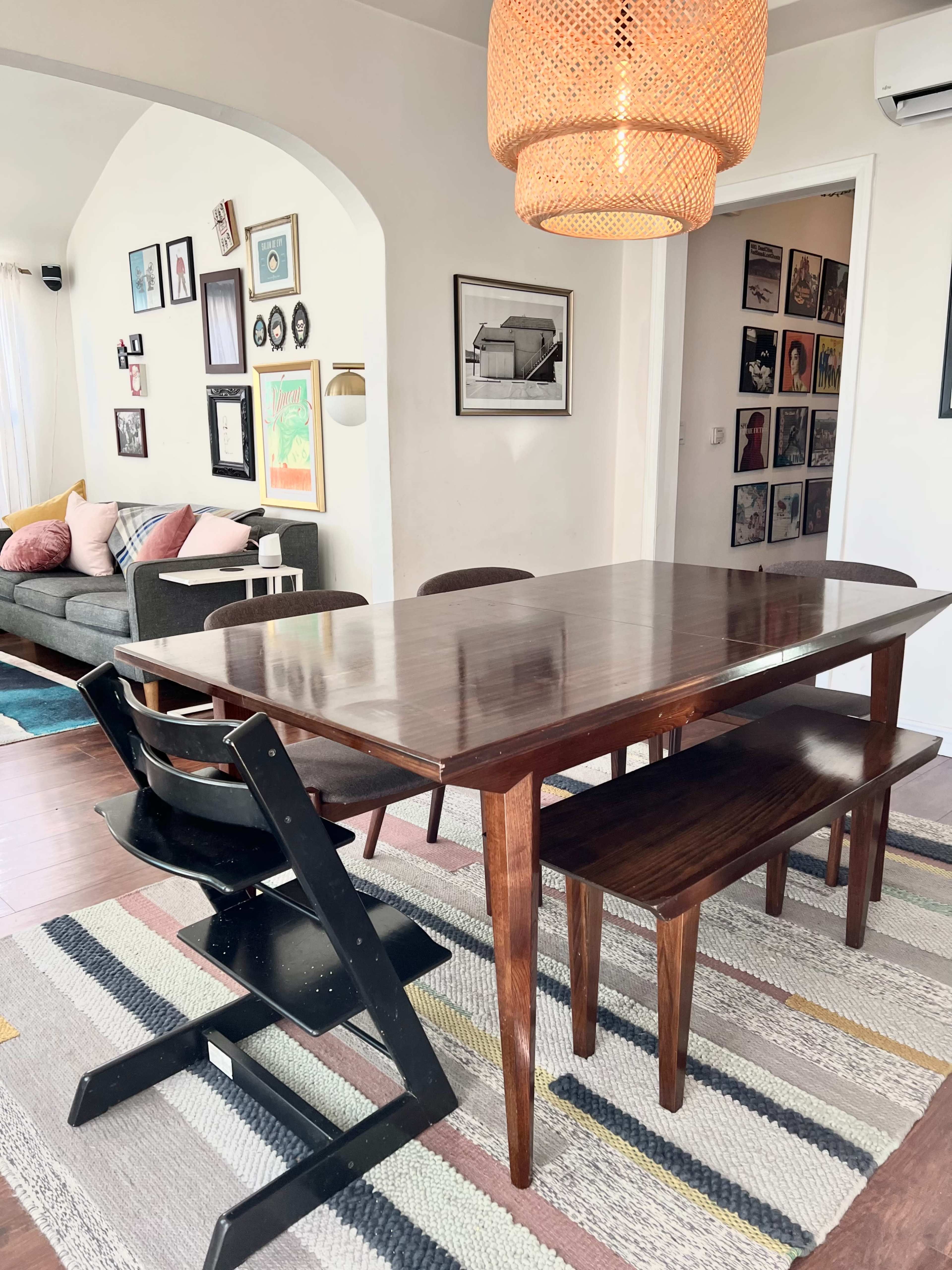 A wooden dining table with a bench and two chairs is positioned under a woven pendant light in a brightly lit room featuring colorful artwork on the walls.