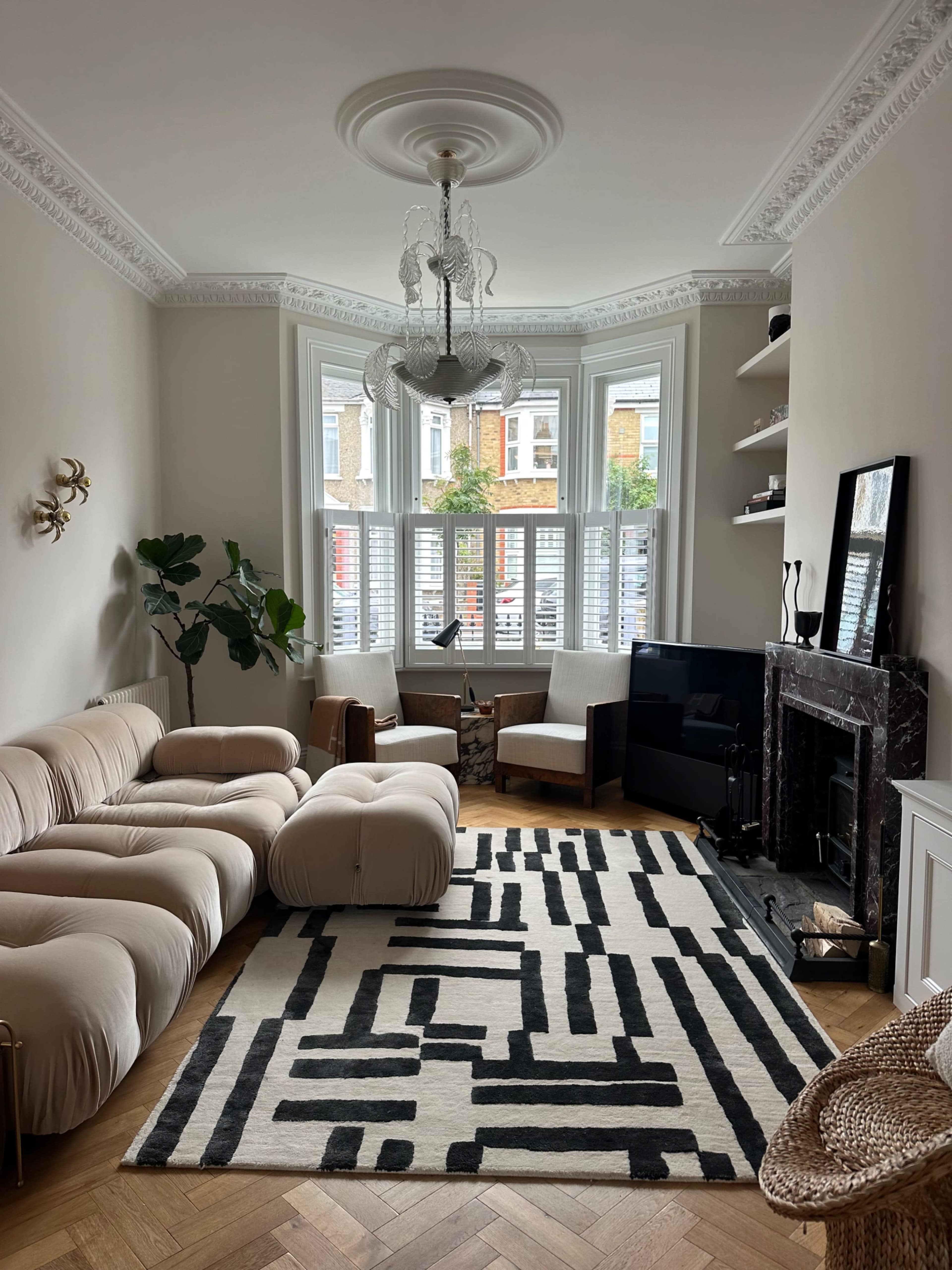 The living room features a curved beige sofa, a patterned black-and-white rug, and large windows with wooden shutters, creating a cozy seating area around a television and a fireplace.
