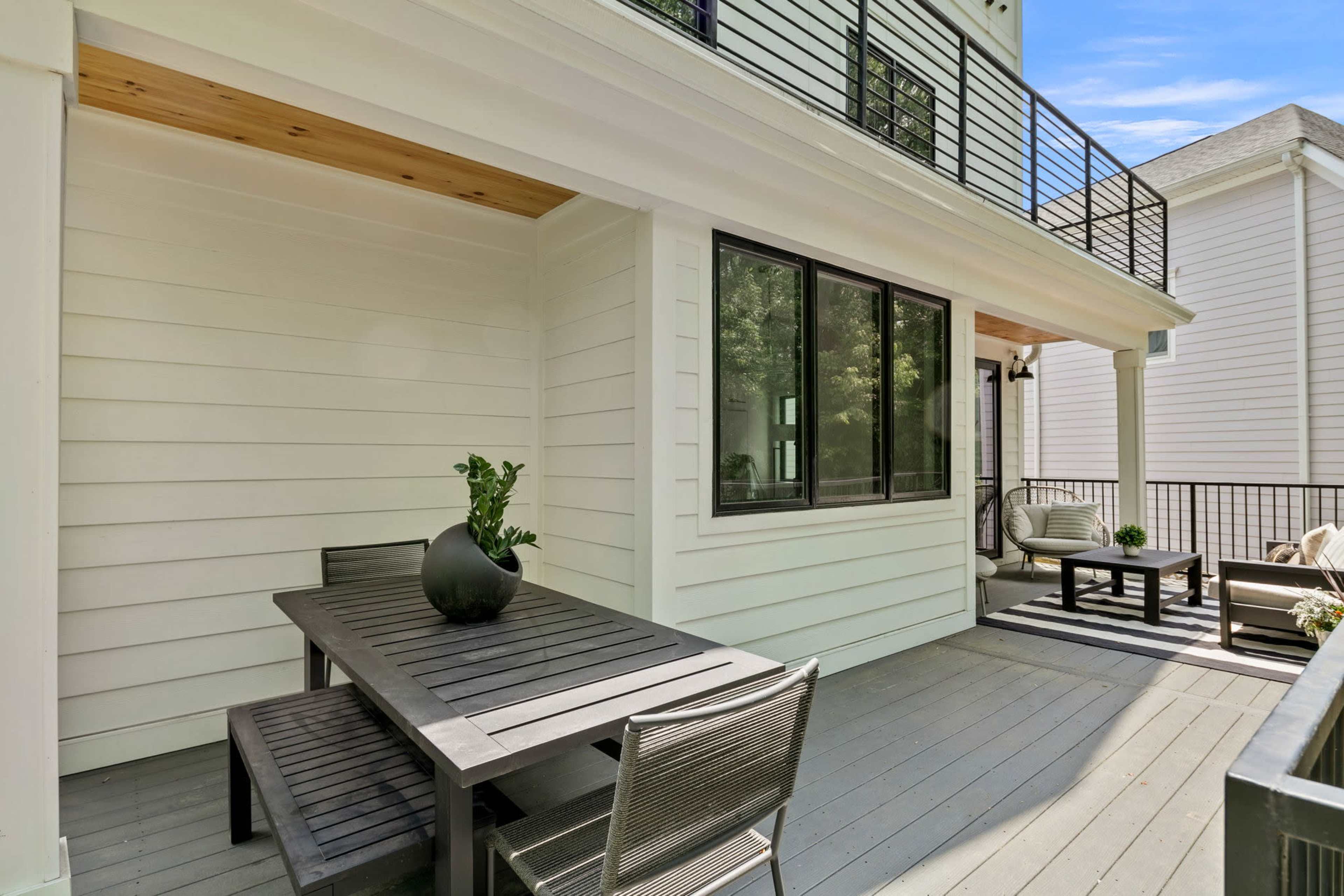 A modern outdoor patio with a wooden table and chairs, surrounded by white siding and a large window.