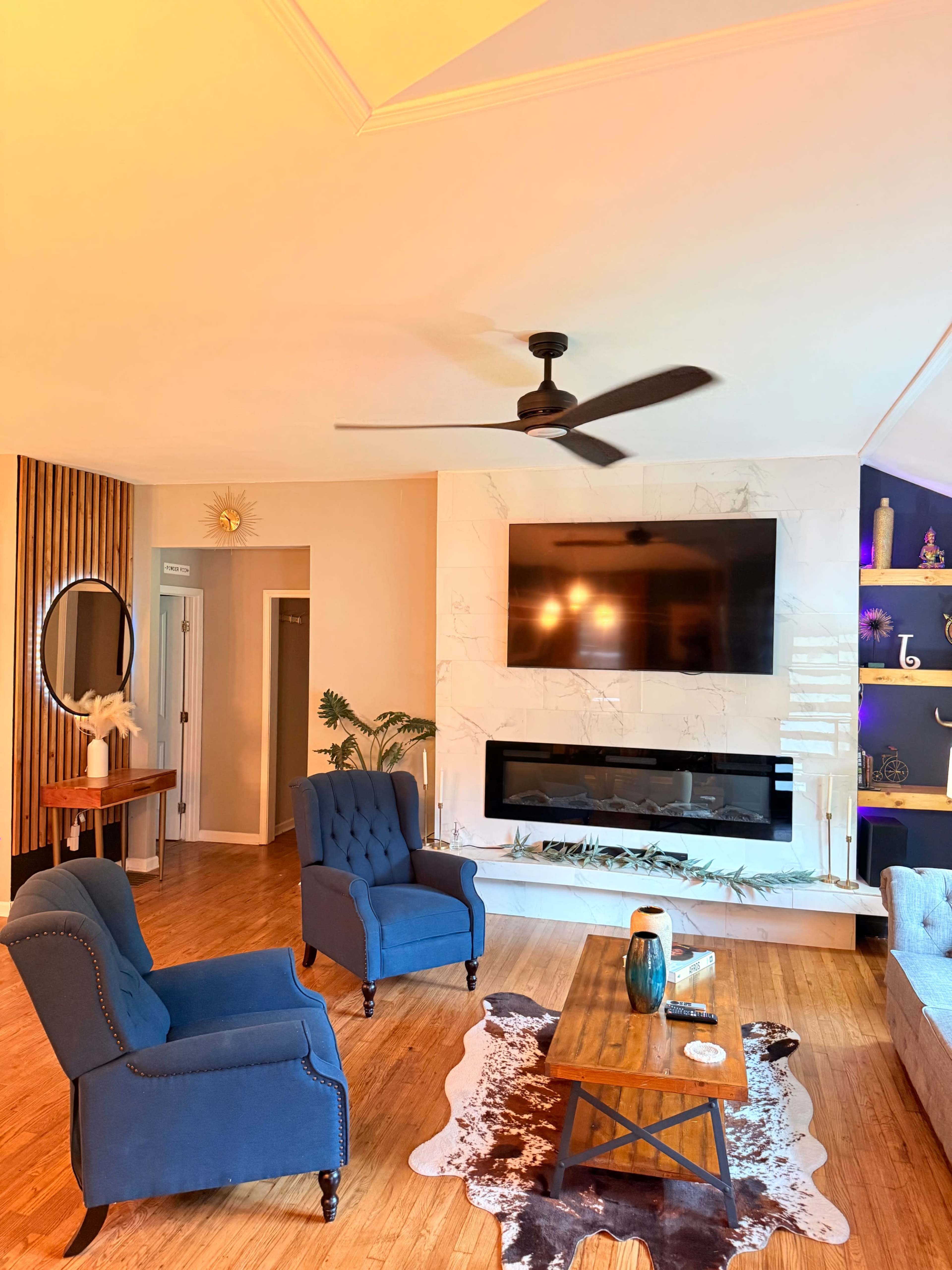 The living room features two blue armchairs, a wooden coffee table, a large wall-mounted TV, and a modern fireplace against a light-colored wall.