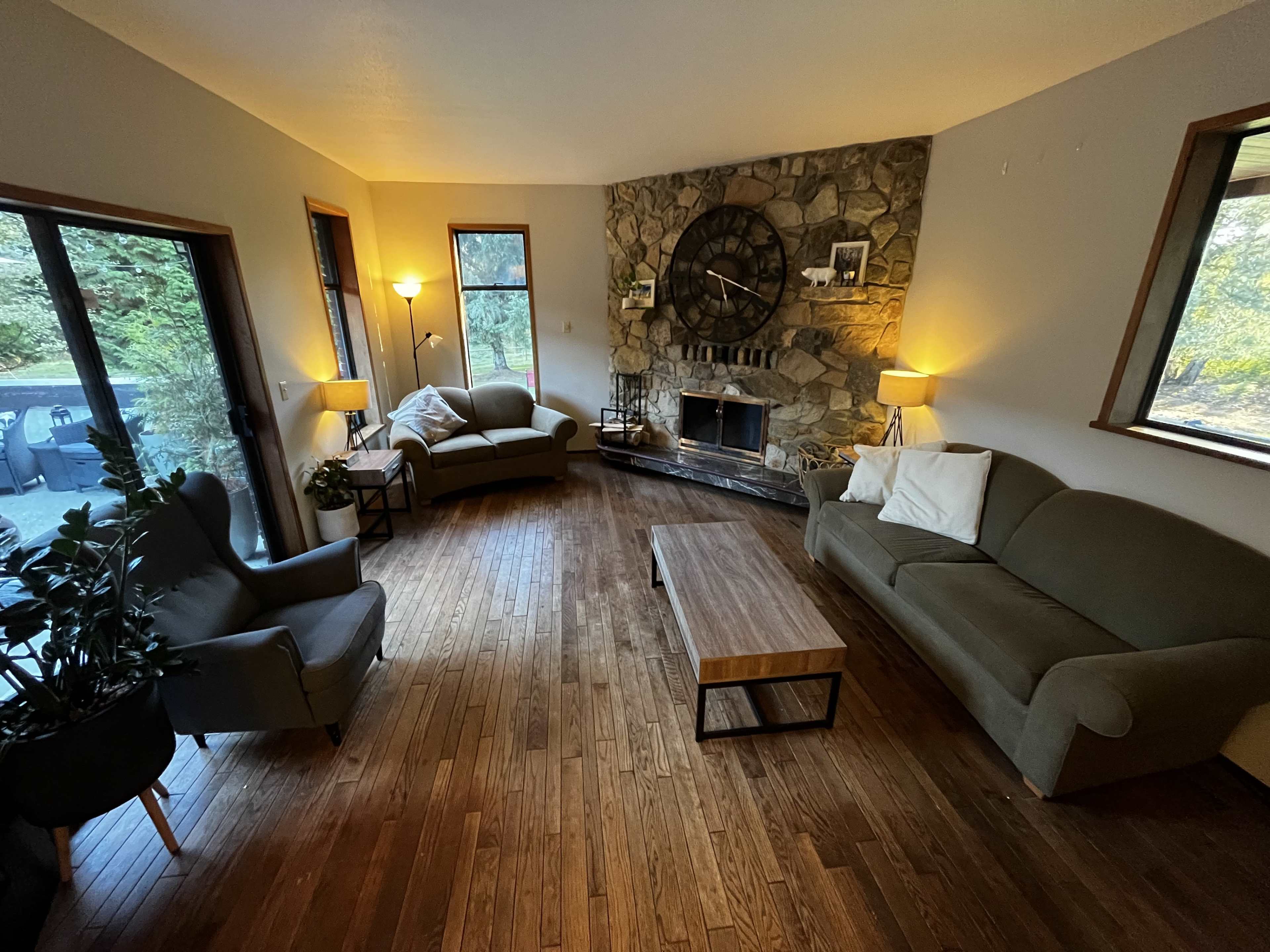 The room features a stone fireplace, two seating areas with green couches, a wooden coffee table, and large windows overlooking a wooded view.