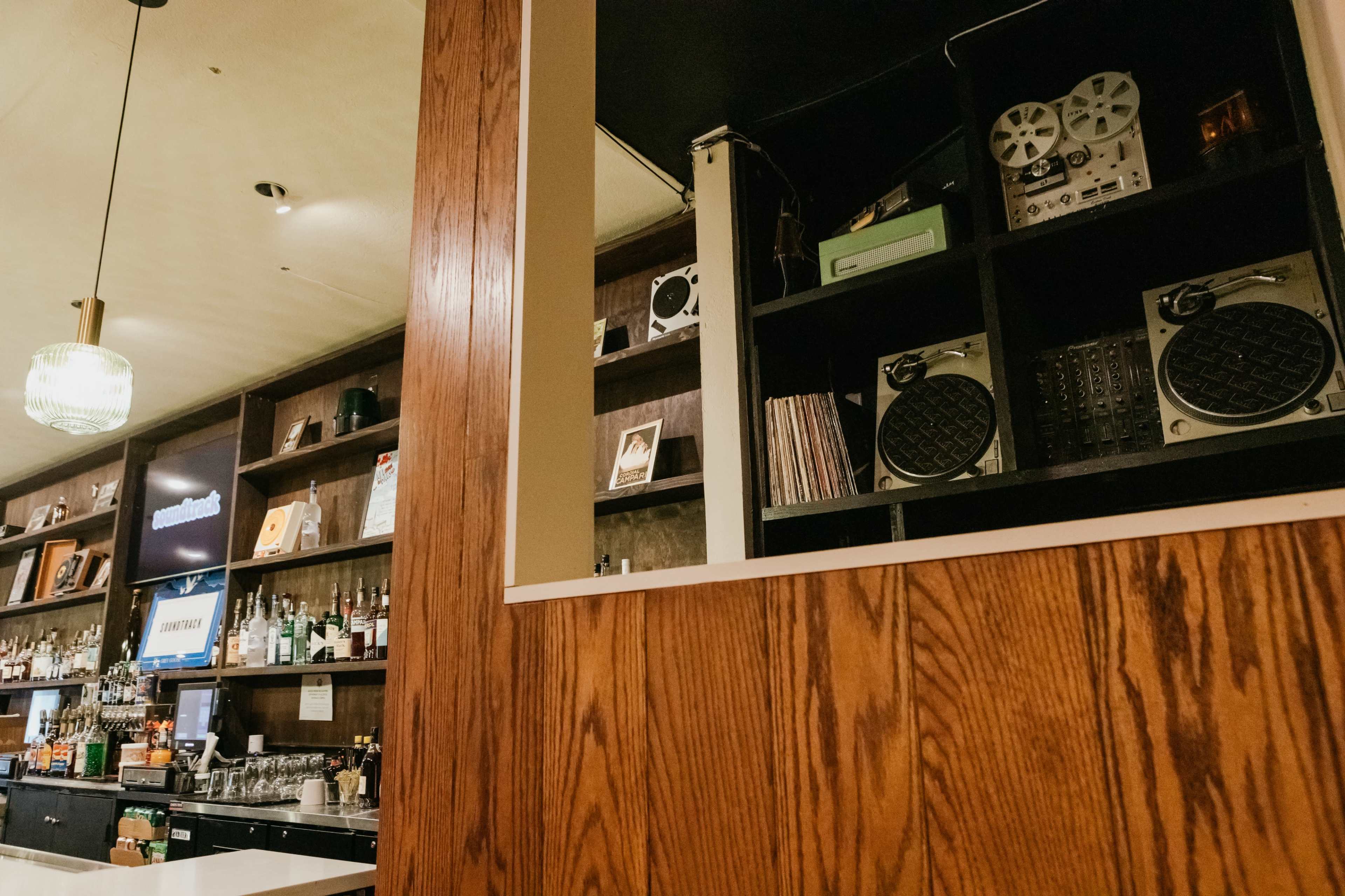 The image shows a bar area with a wooden partition in the foreground and various audio equipment, including speakers and vinyl records, displayed on shelves in the background.