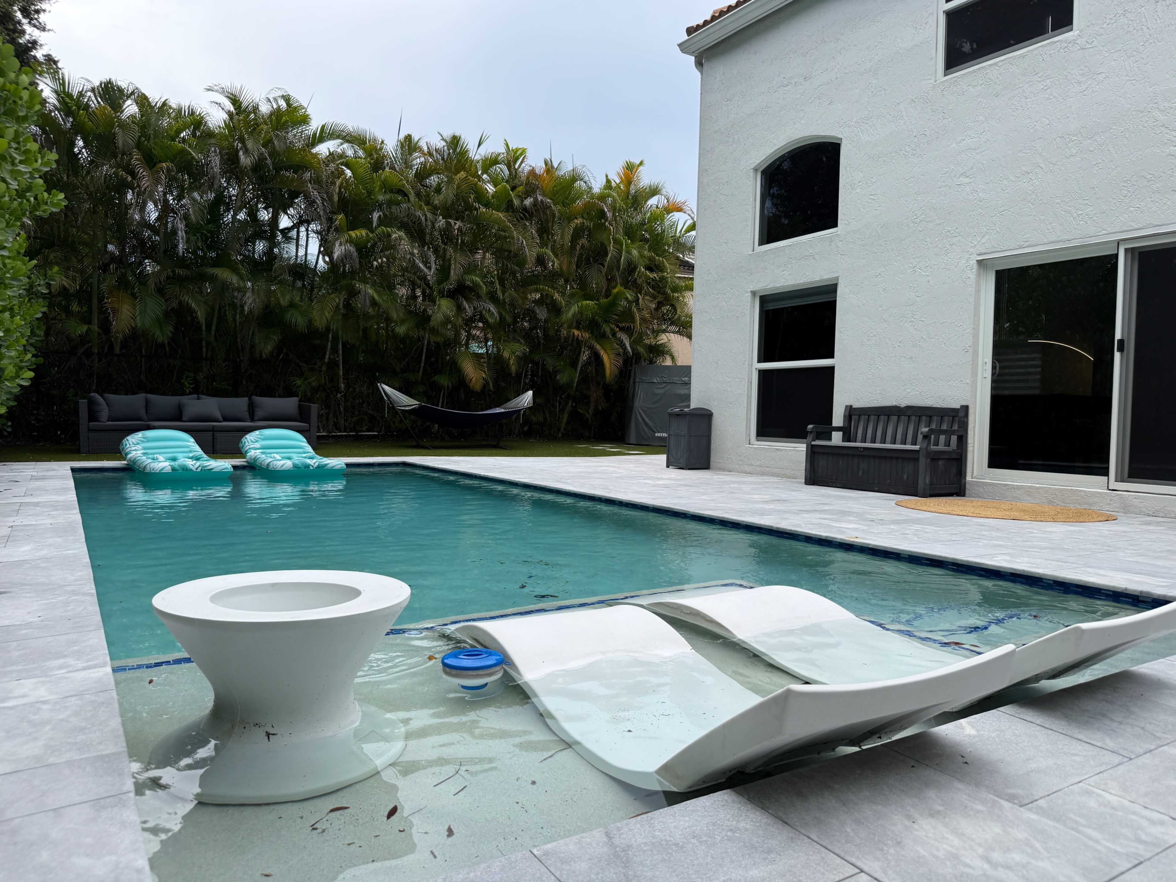 The image shows a pool area with two lounge chairs and a small table beside the water, surrounded by greenery and a house with large windows.