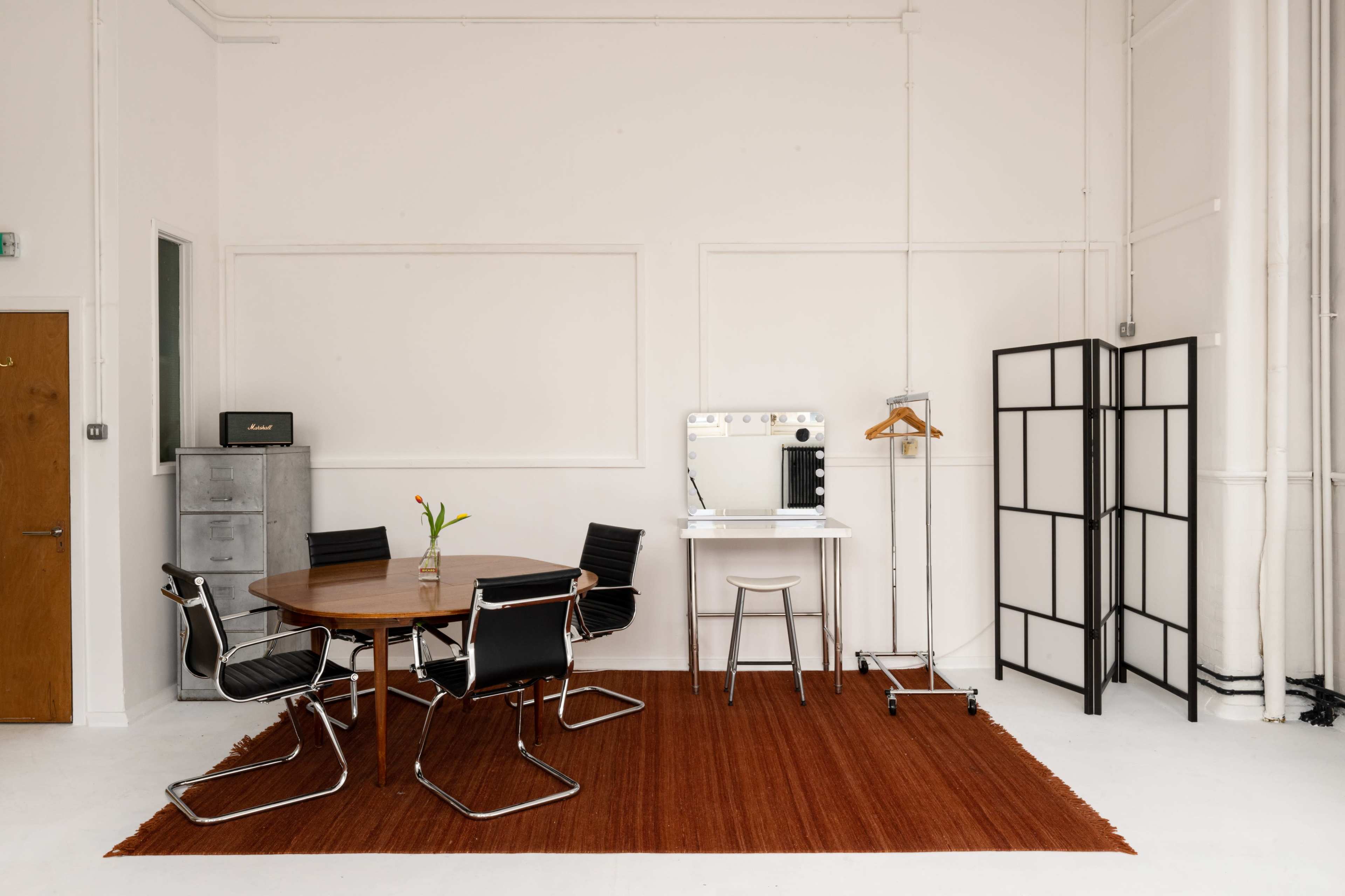 The image shows a minimalist room with a round wooden table and black chairs, a vanity mirror with lights, a clothing rack, a folding screen, and a reddish rug on the floor.