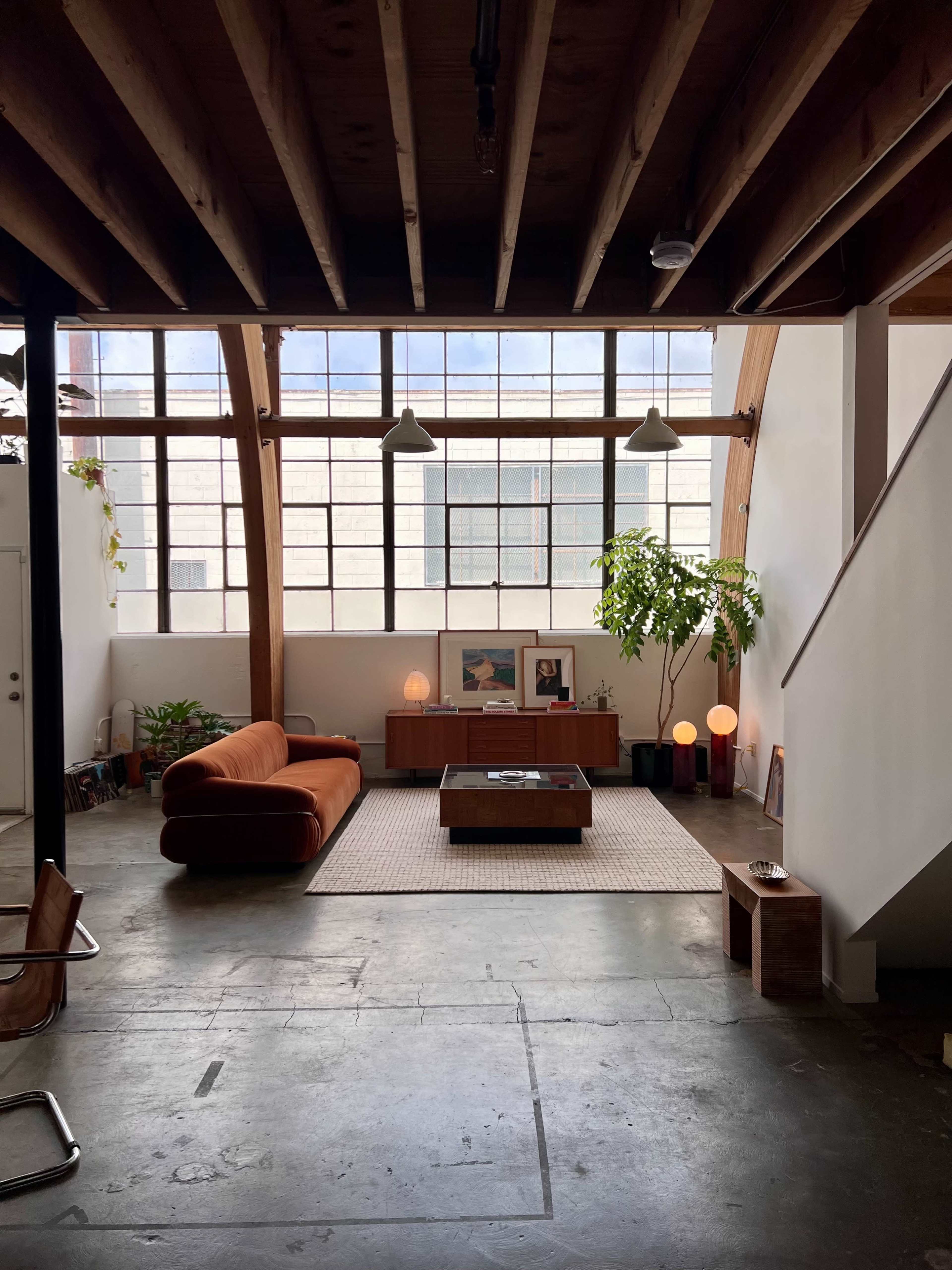 A large, open living space featuring a brown sofa, a wooden coffee table, and a wall of tall windows with a view of the outside, complemented by plants and artistic decor.