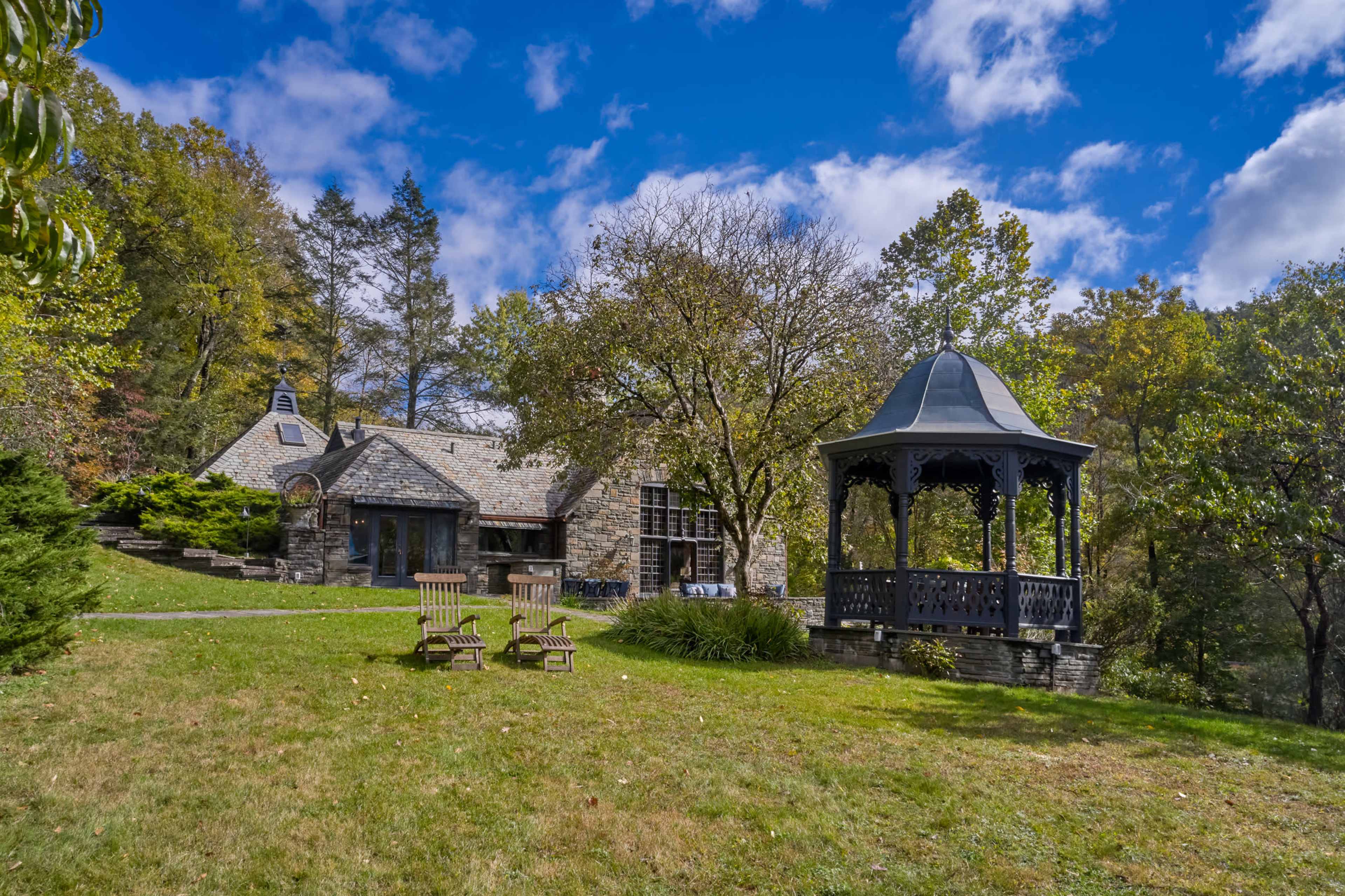 The image depicts a stone house beside a gazebo and two wooden chairs on a grassy lawn surrounded by trees under a partly cloudy sky.