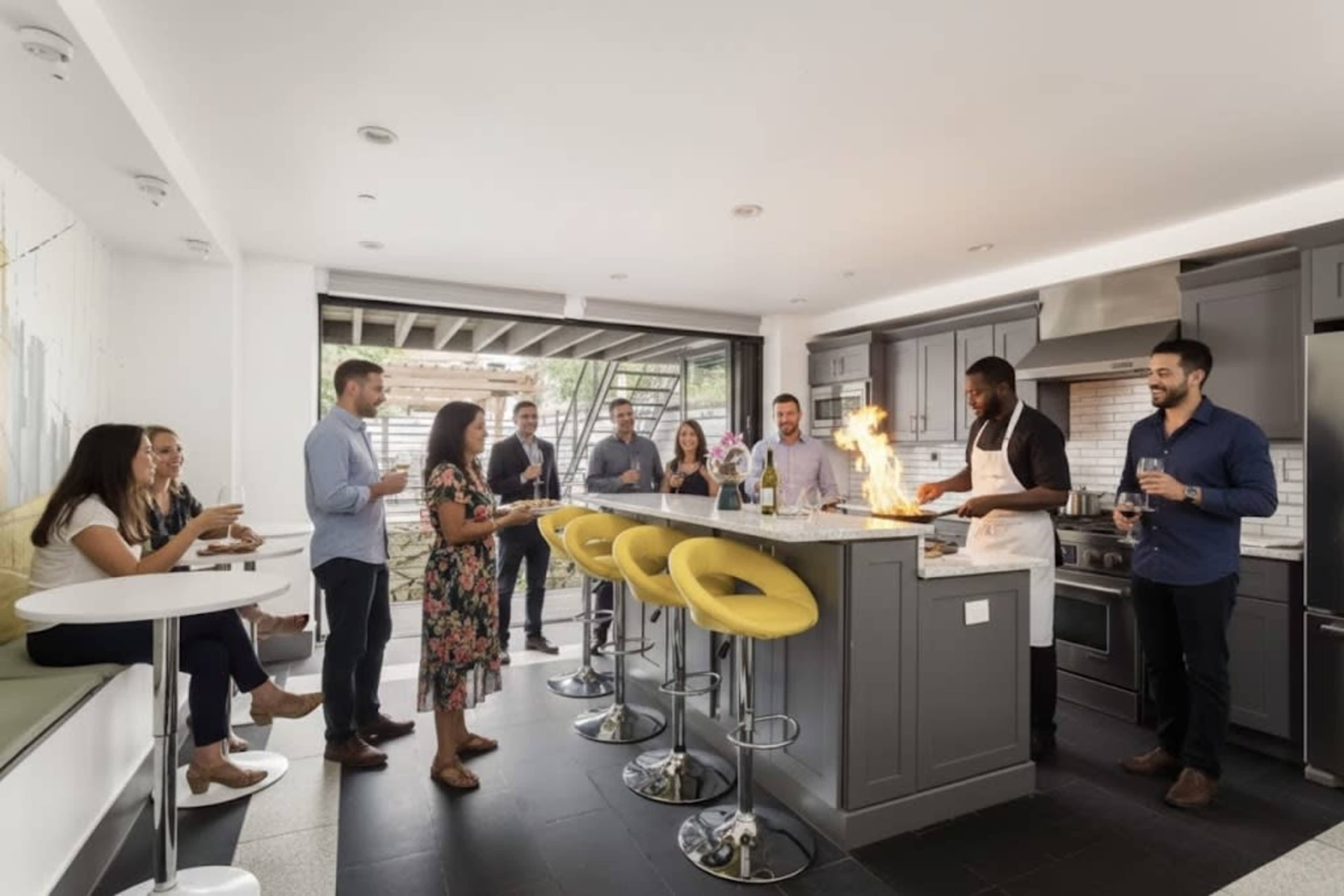 A group of people gathers in a modern kitchen as one individual cooks and flames rise from the stovetop.