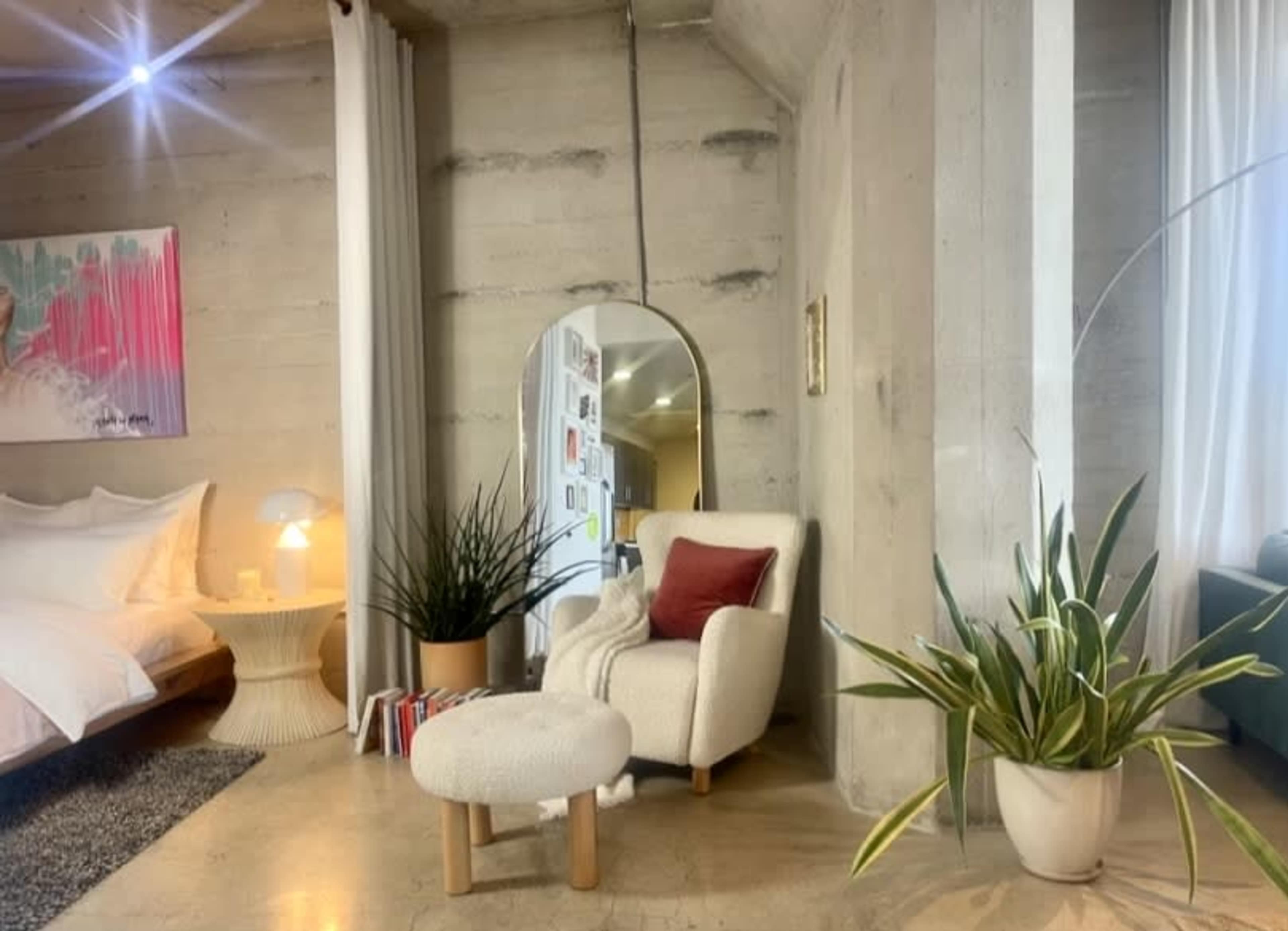 The image shows a modern bedroom setup with a bed, decorative plants, an armchair, and a large mirror against a concrete wall.