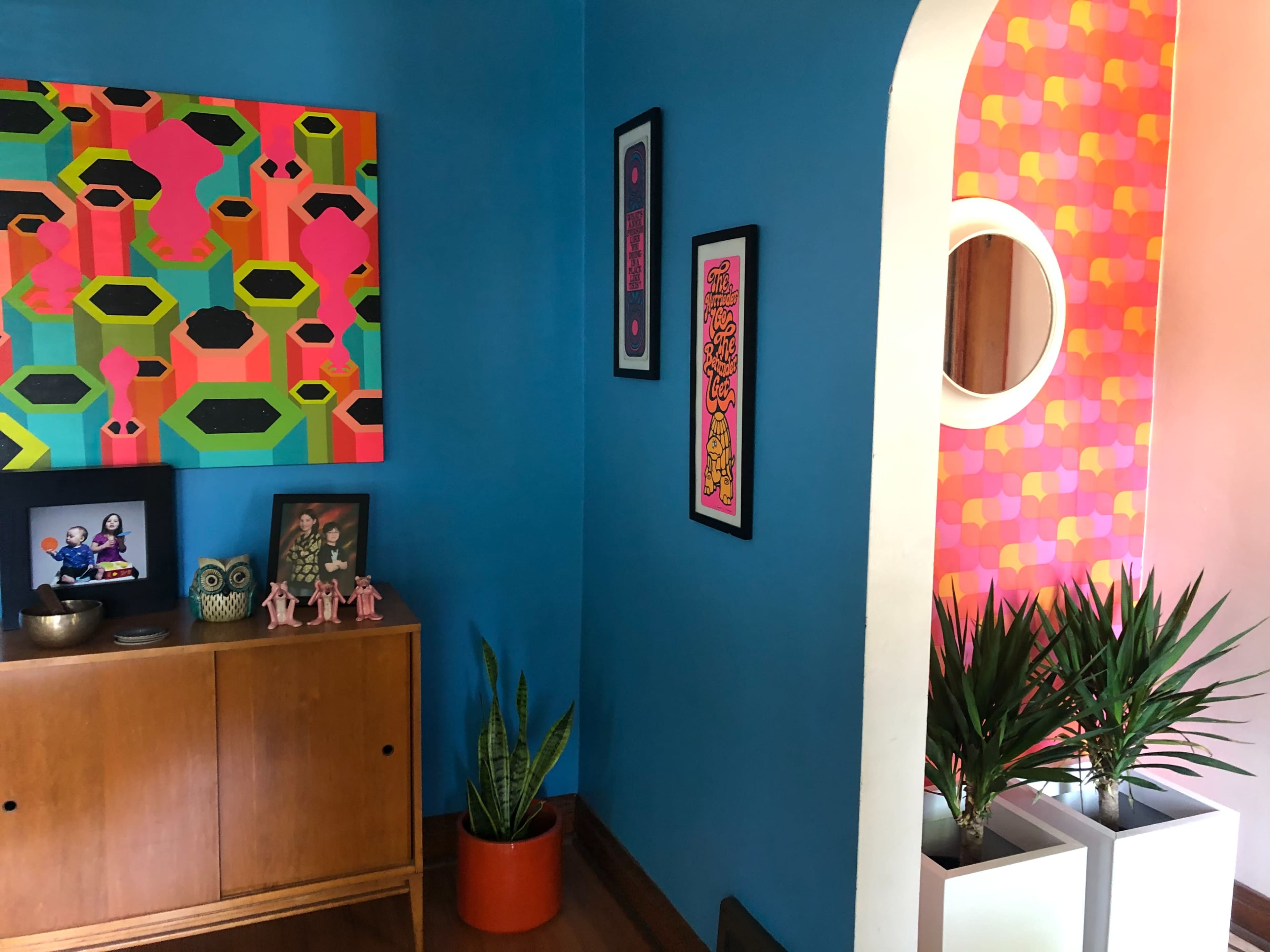 The image shows a vibrant room with blue walls, colorful artwork, a wooden cabinet with decorative items, and plants in white planters.