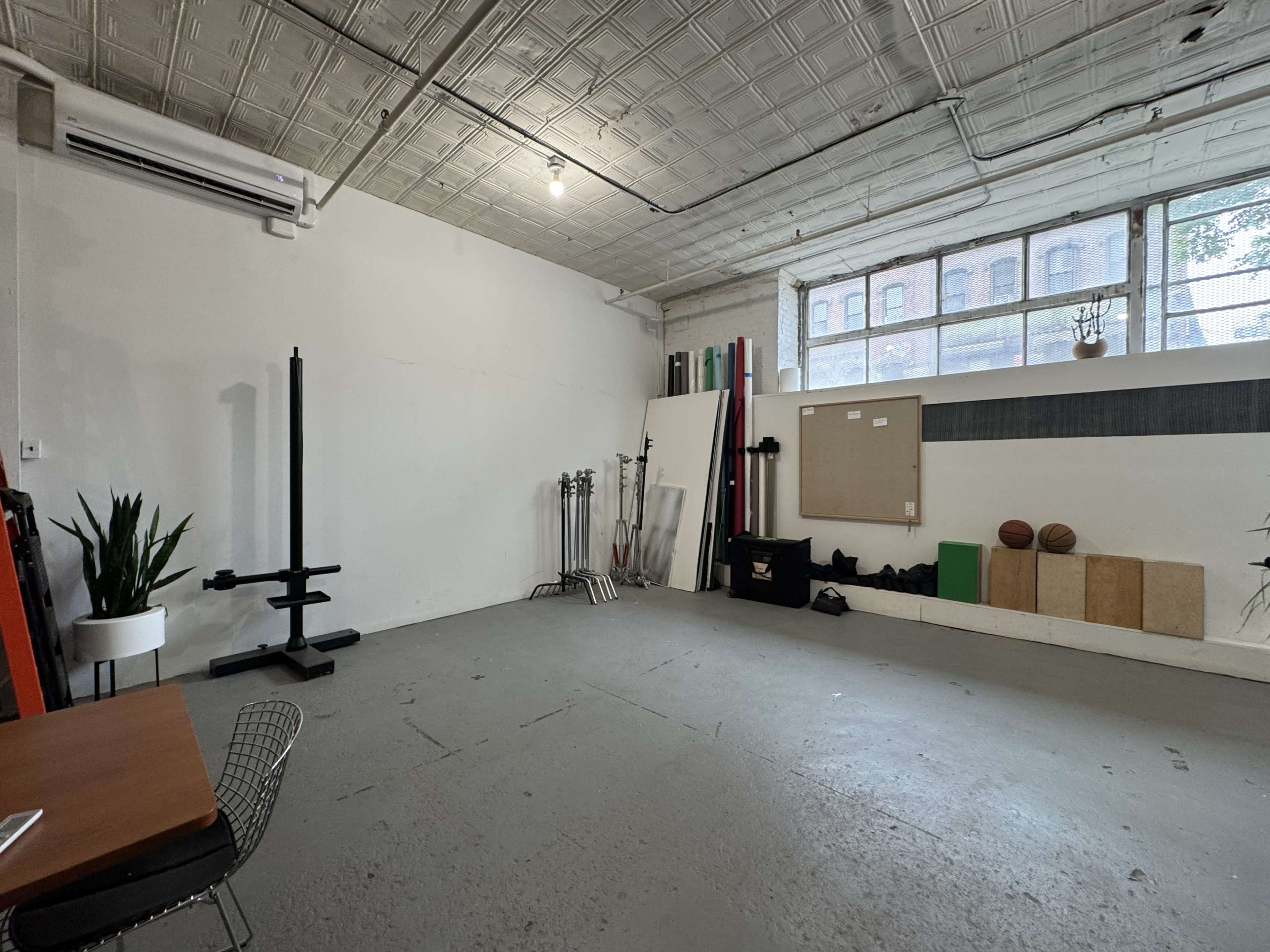 The image shows a spacious, undecorated room with a concrete floor, featuring gym equipment, exercise mats, and a table with a chair against one wall.