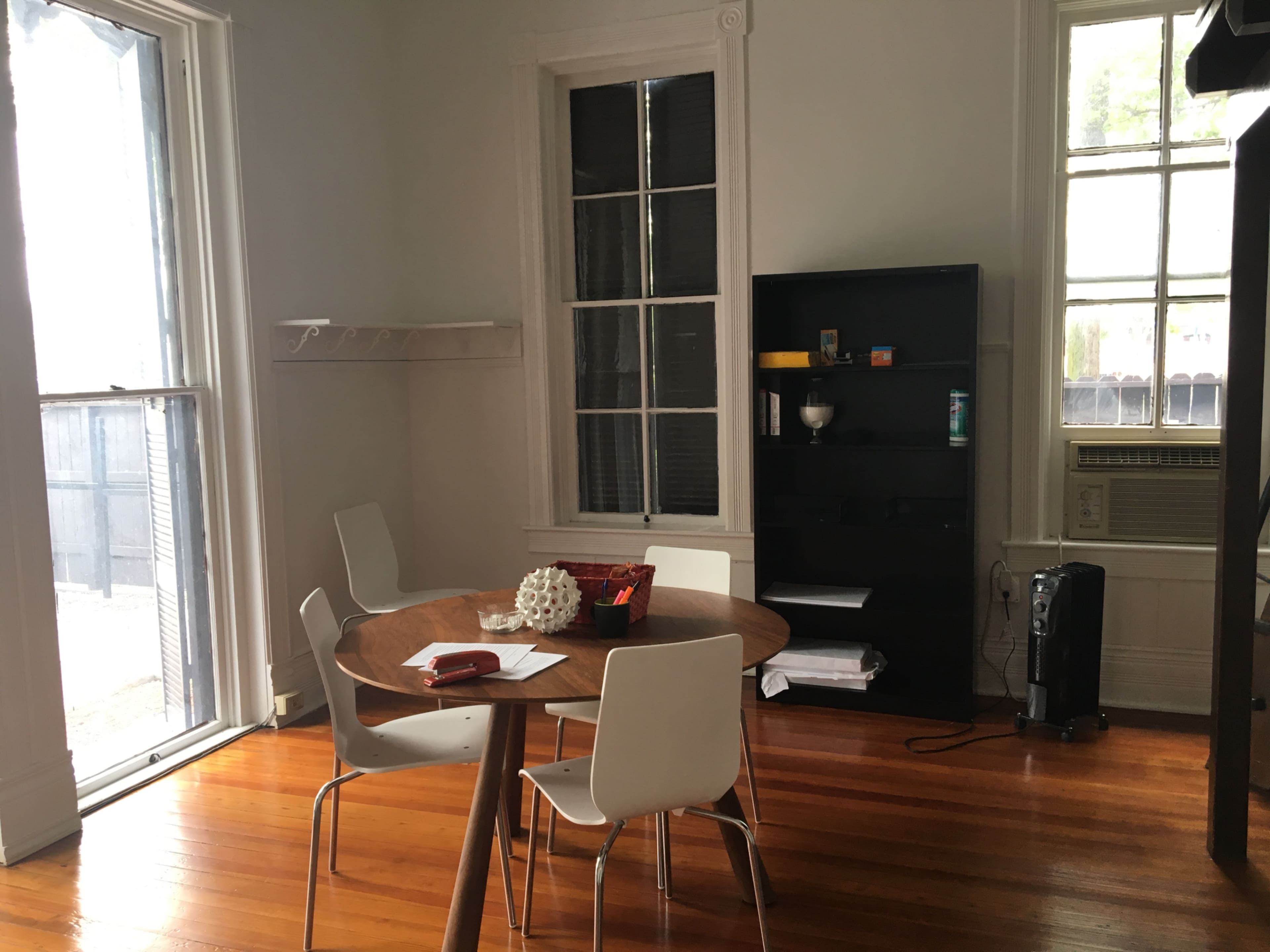 A bright room with a round wood dining table surrounded by white chairs, a black shelving unit, and a window with a view of the outdoors.