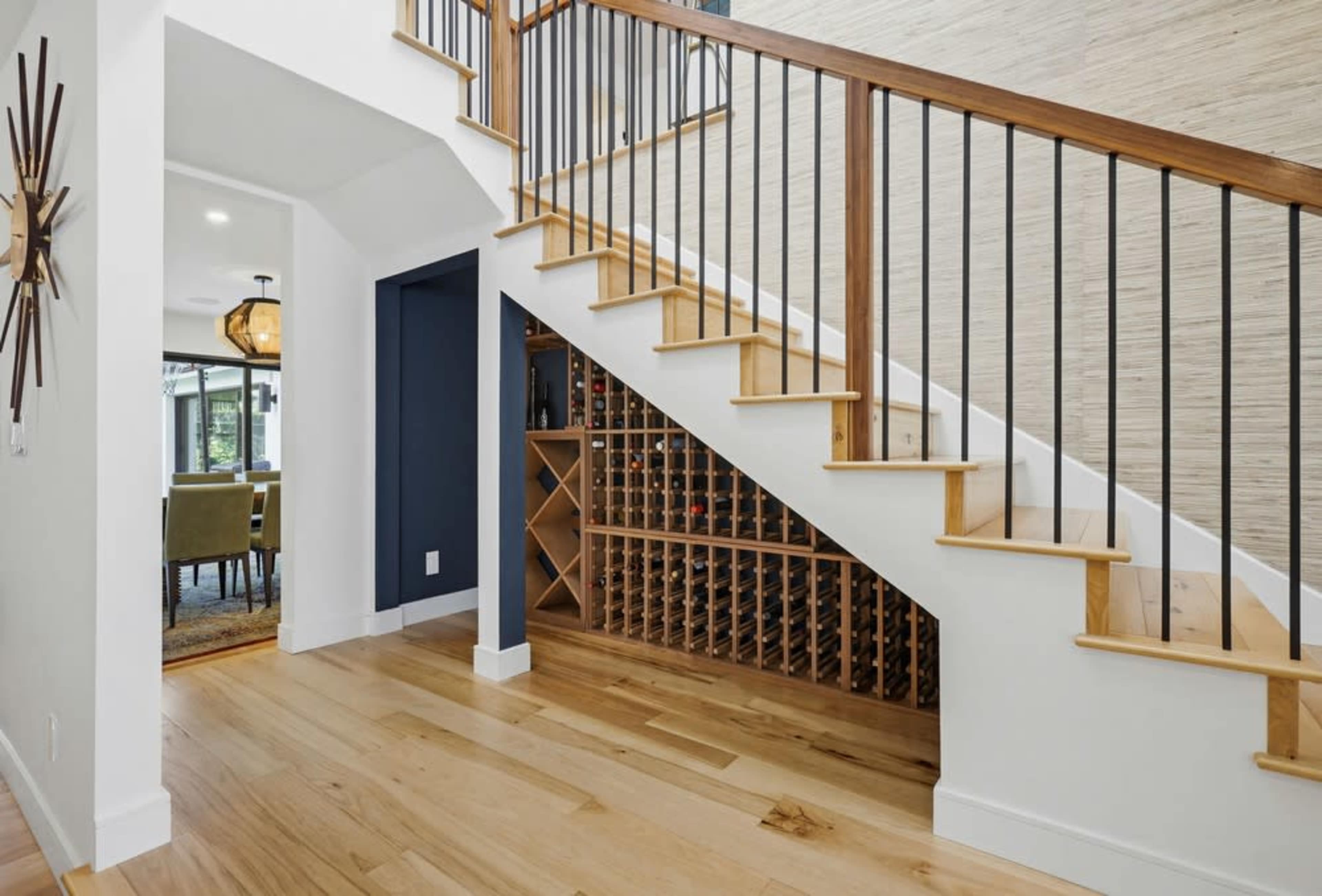 The image shows a modern entryway with a staircase, featuring wooden steps and metal railings, alongside a wine storage unit beneath the stairs.