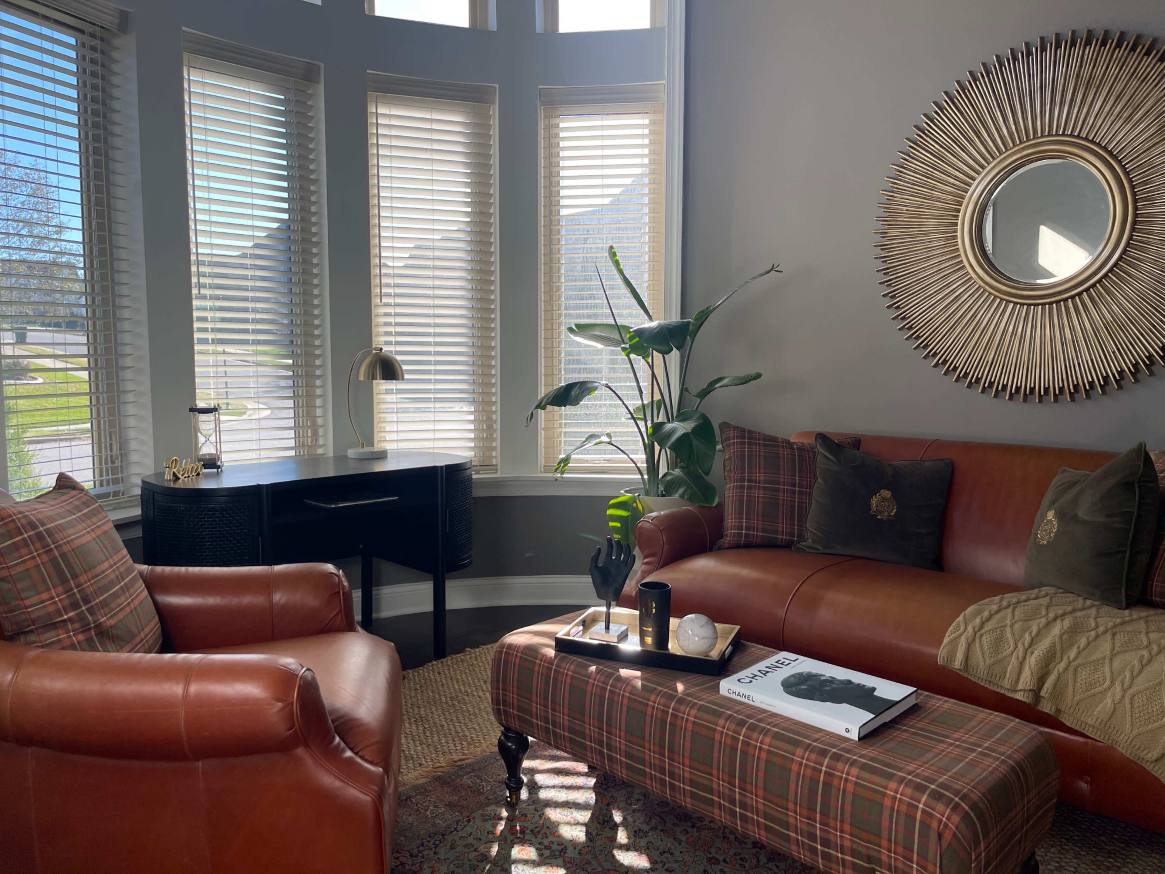 The image shows a cozy living room with a leather sofa, a plaid armchair, a coffee table with decor, and large windows allowing natural light to enter.