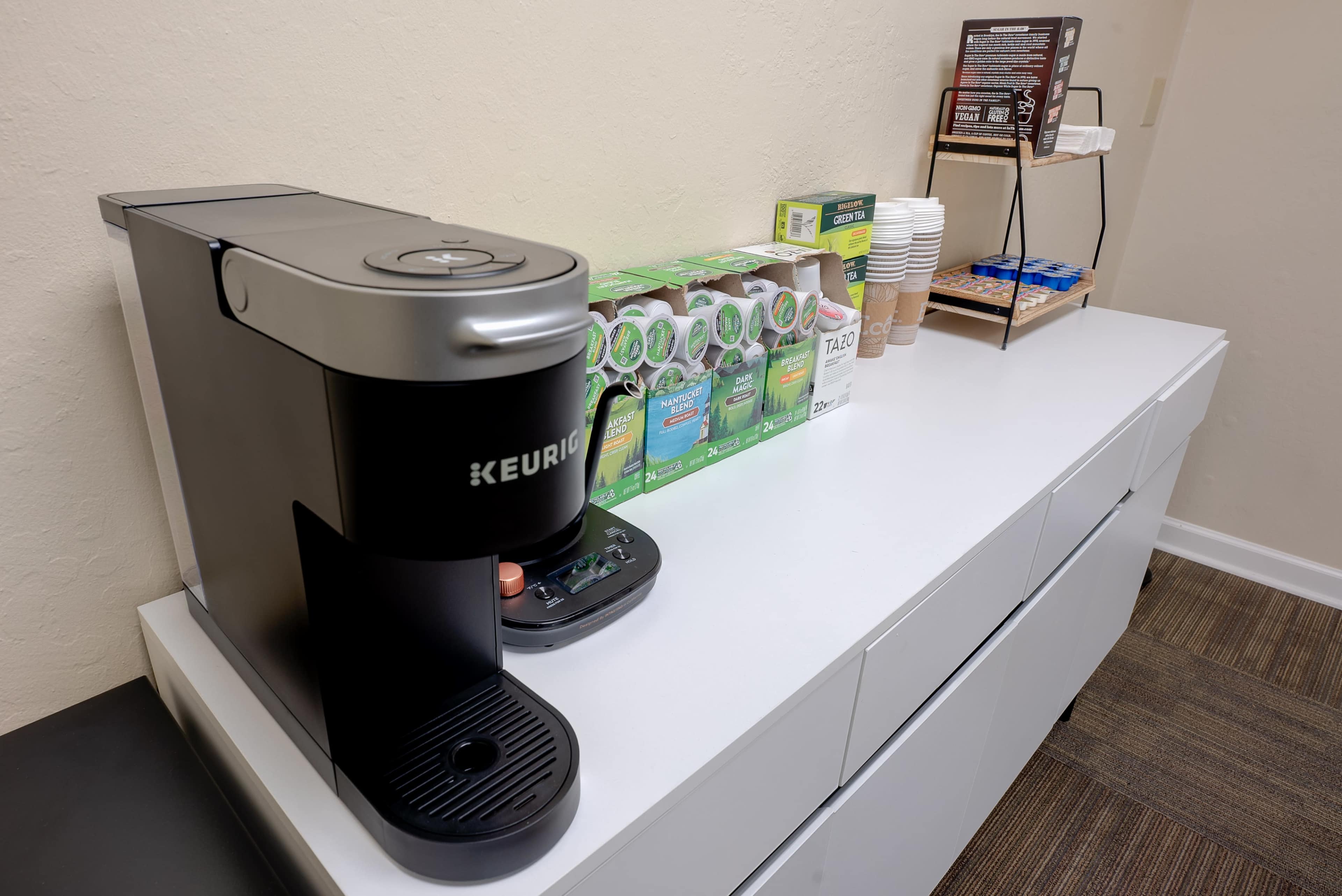 The image shows a Keurig coffee maker on a white counter with a selection of coffee pods, cups, and condiments arranged nearby.