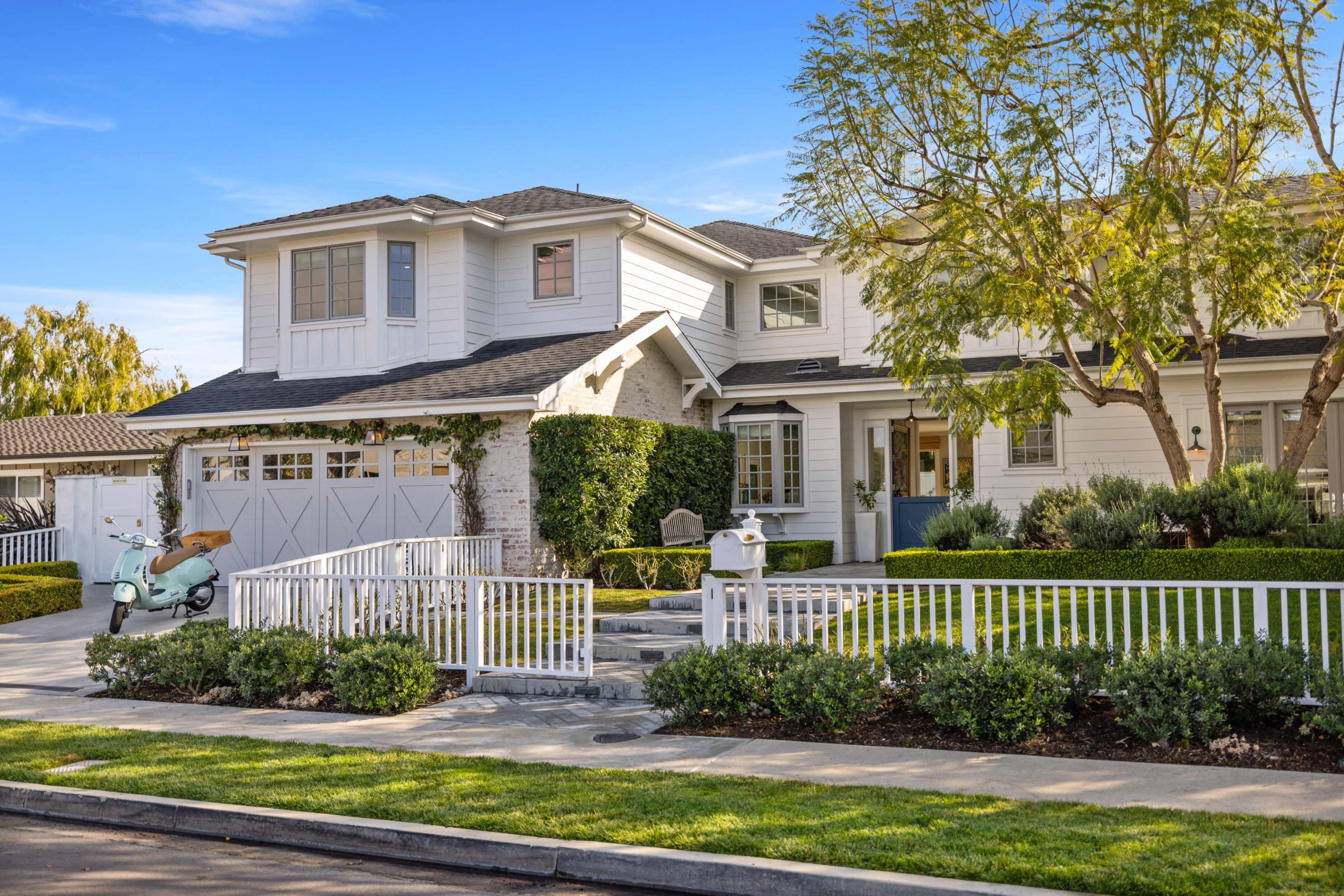 A large, two-story white house with a stone facade features a front porch, greenery, and a vintage scooter parked near a white picket fence.