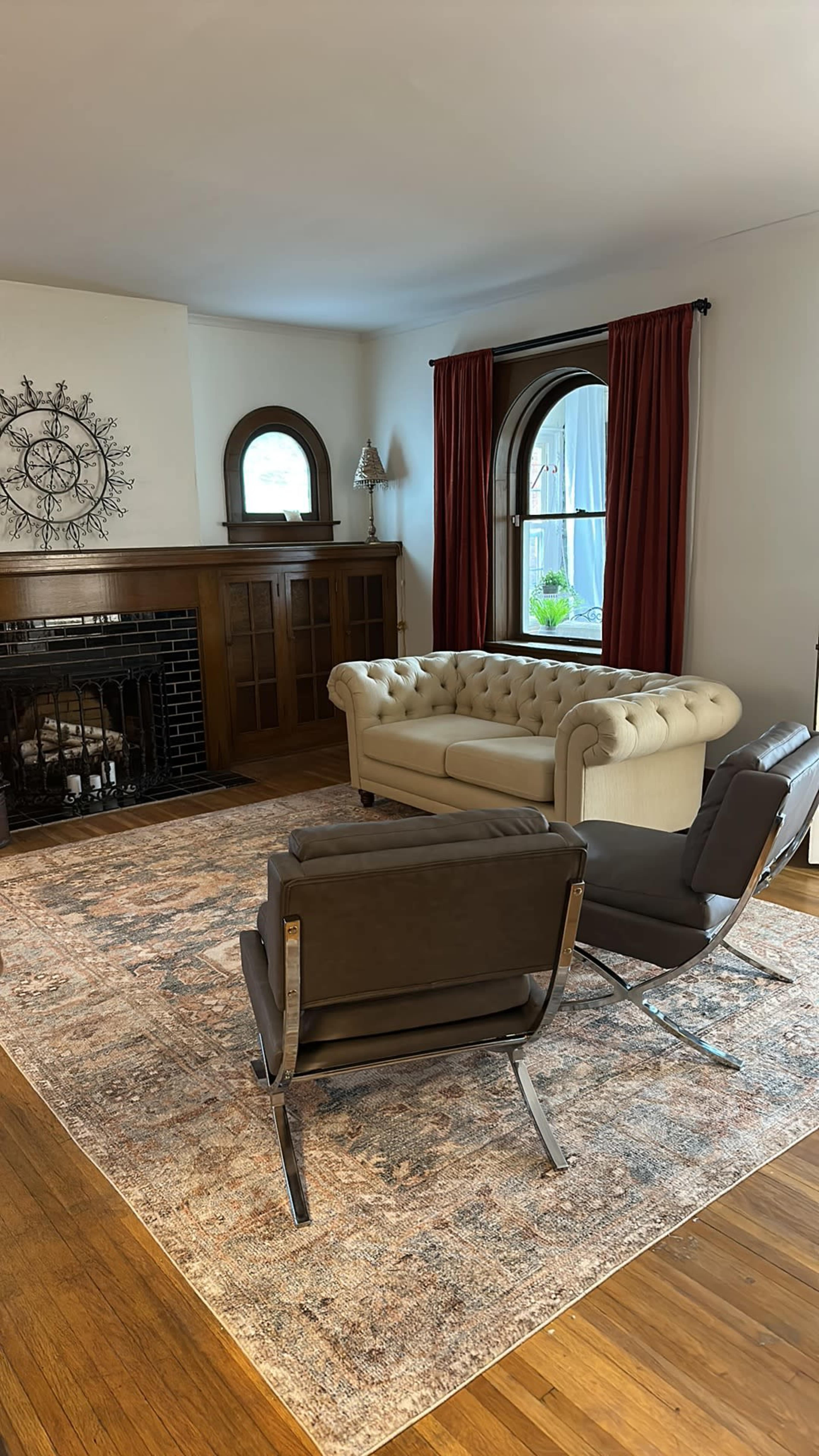 A light-colored tufted sofa sits in a living room with hardwood floors, accompanied by two modern chairs and a large area rug.