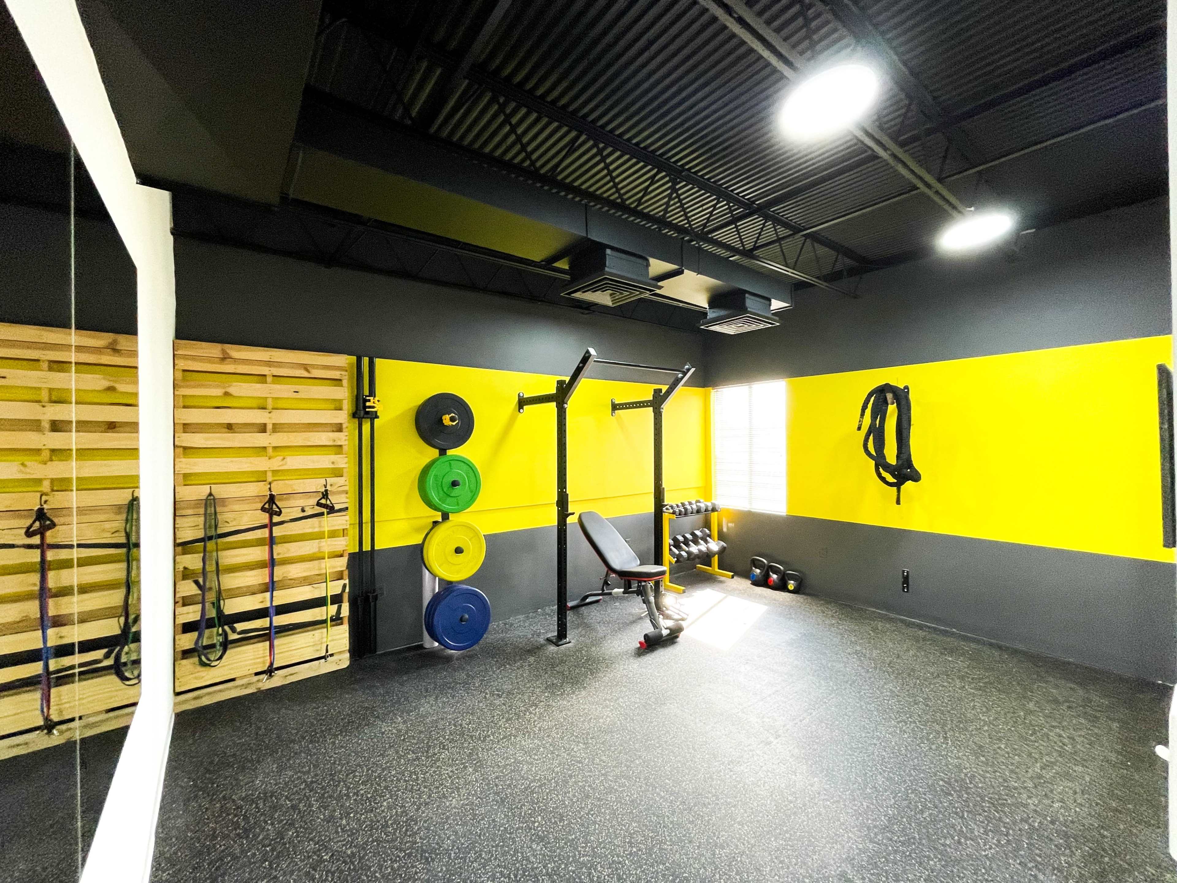 The image shows a gym interior with weightlifting equipment, including a squat rack, dumbbells, and resistance bands against a yellow and gray wall.