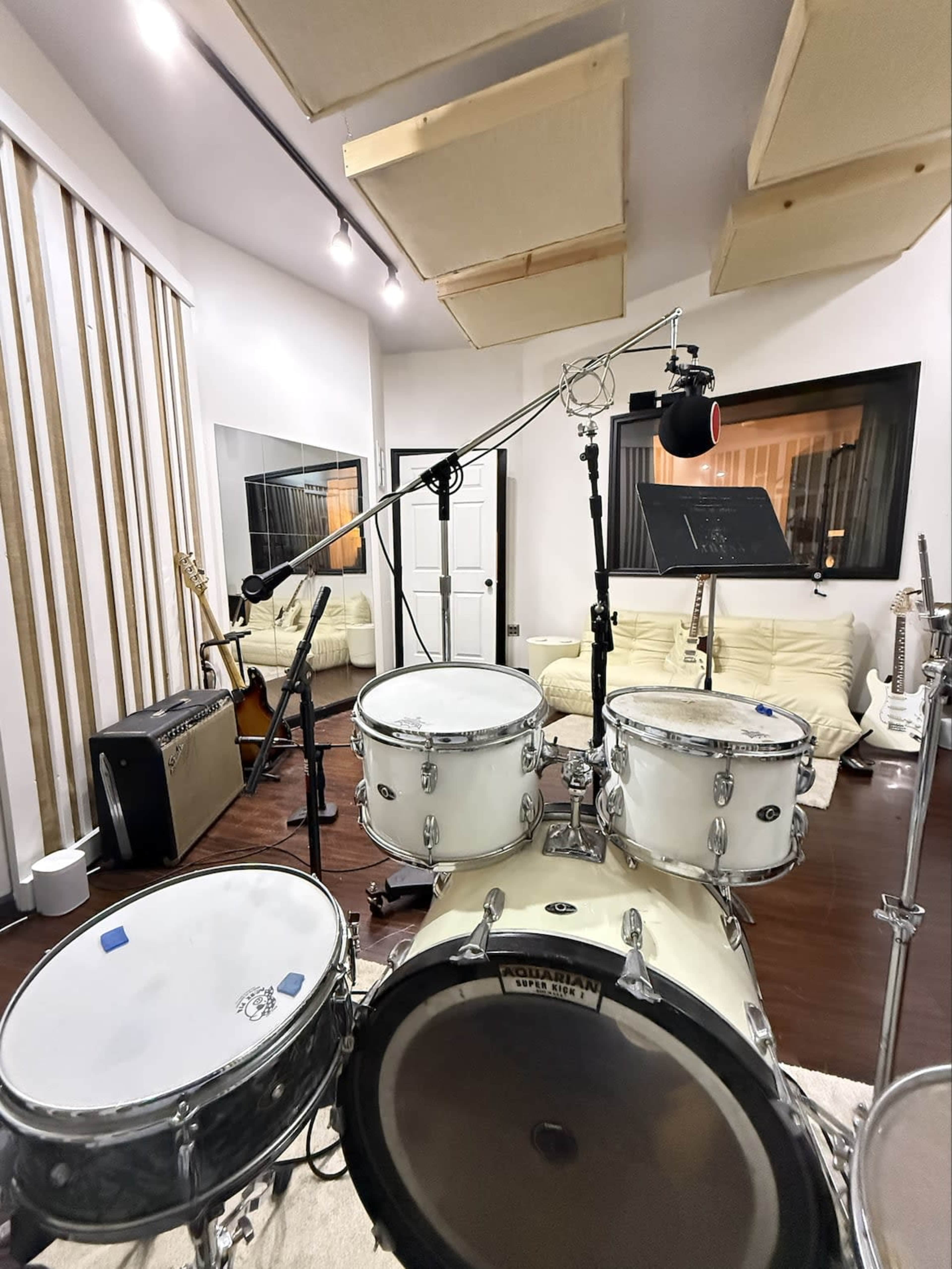 A drum set is positioned in a music studio with soundproofing panels, a microphone on a stand, and various instruments visible in the background.