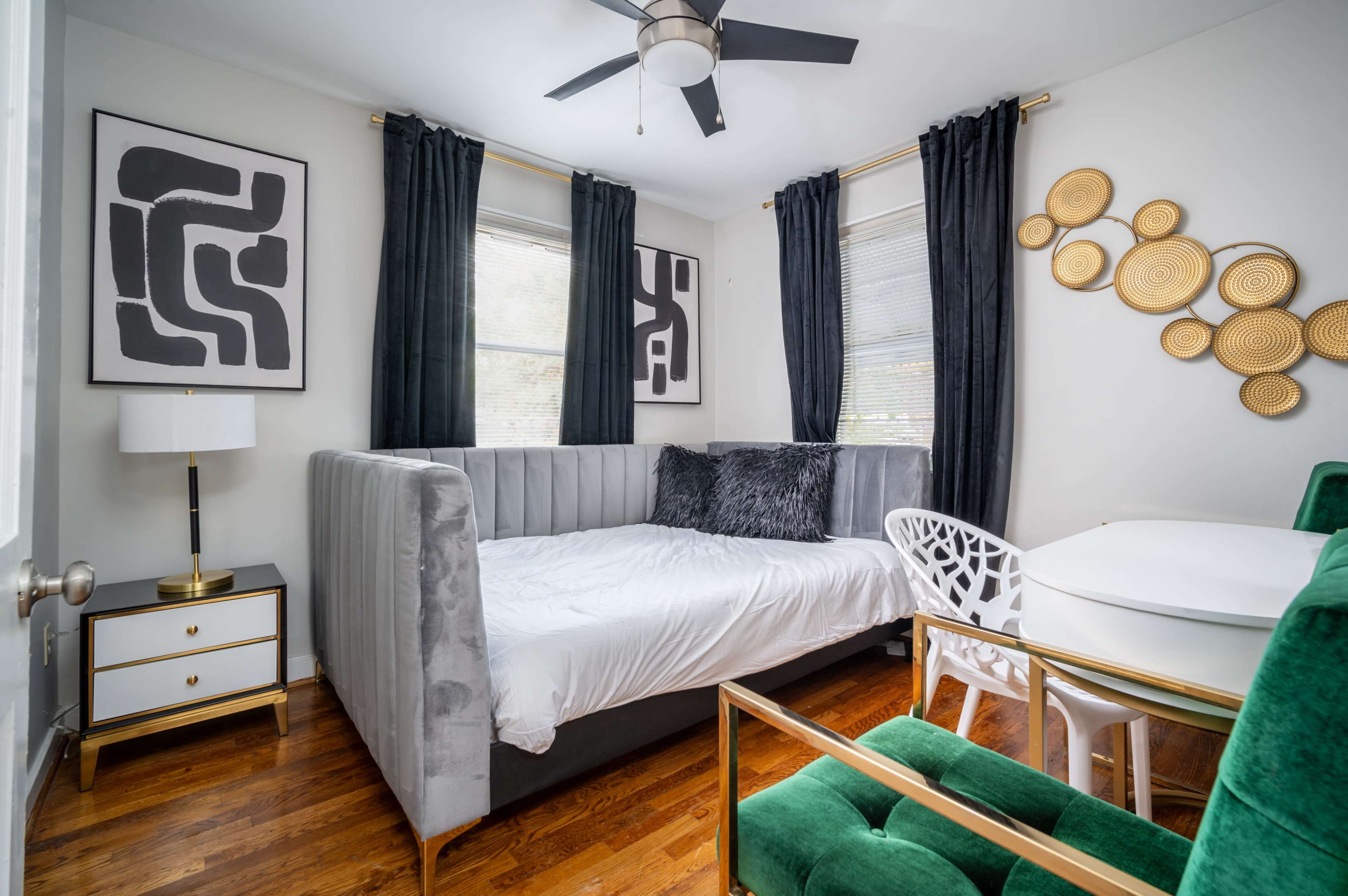 A modern bedroom with a grey upholstered daybed, black curtains, abstract wall art, and a green chair next to a small dining table.