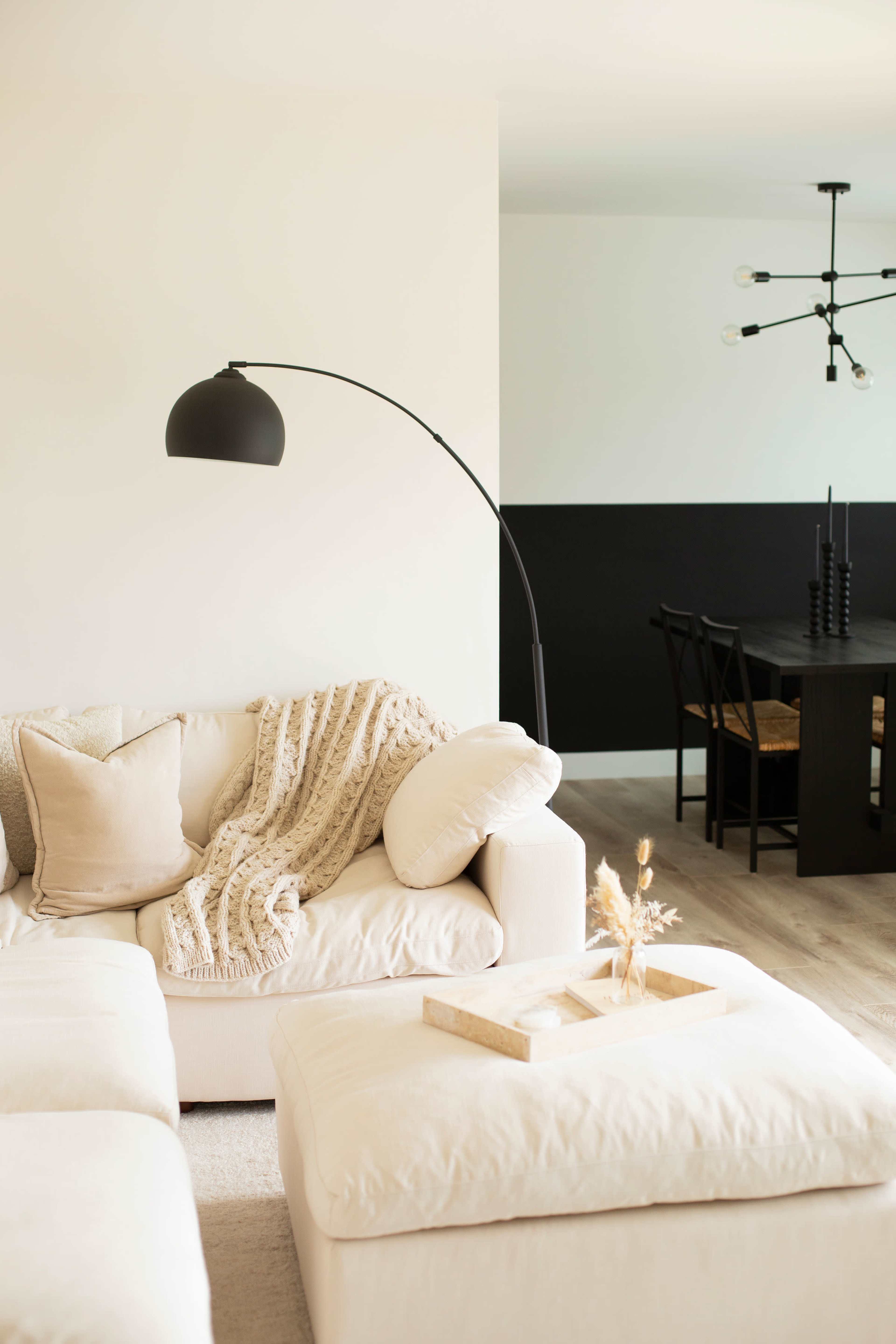 A cozy living room features a beige sectional sofa adorned with a textured throw and a modern arc floor lamp, alongside a minimalist dining area with a black accent wall and a wooden table.