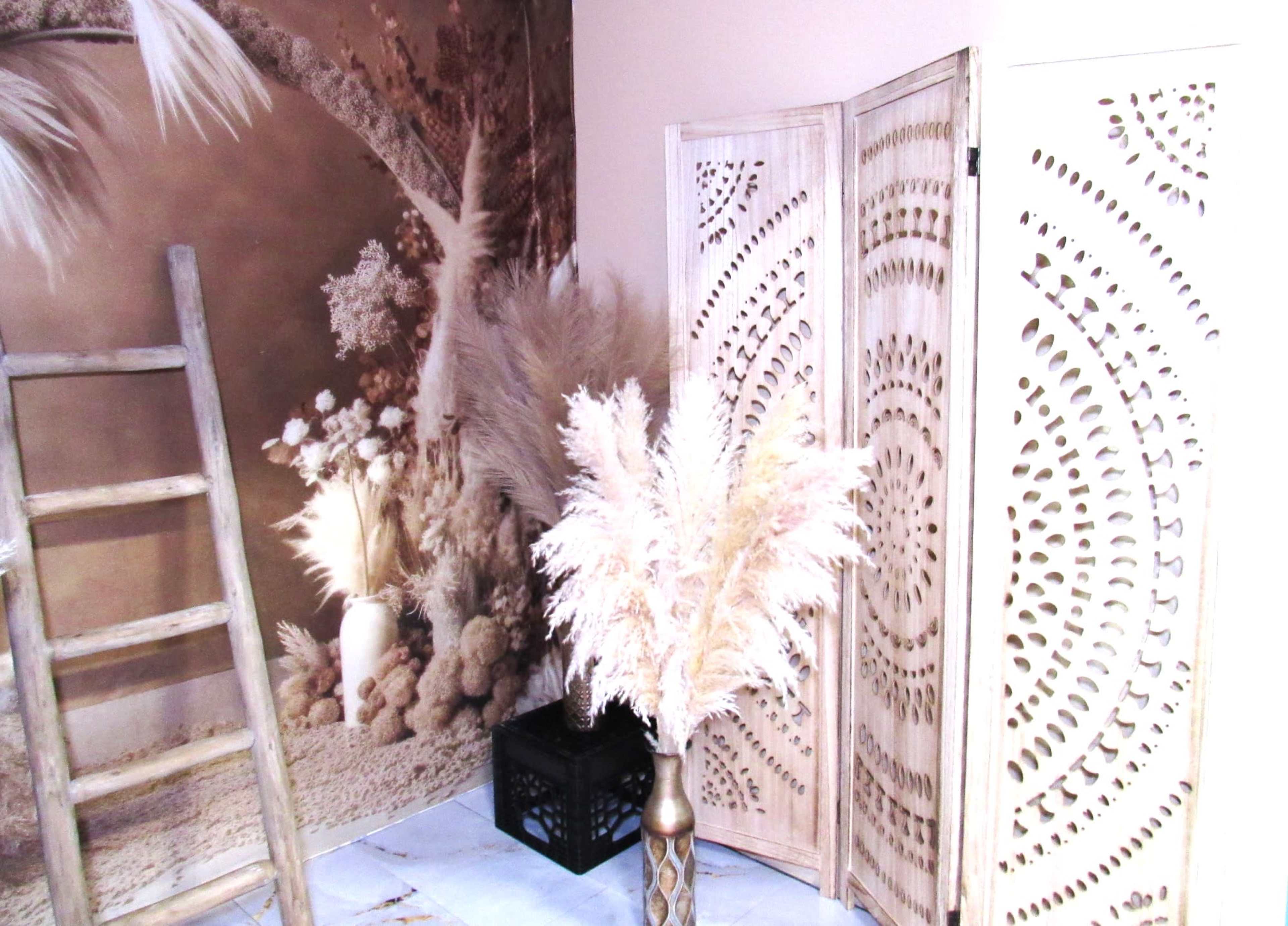 A corner of a room with a decorative folding screen, a vase of dried pampas grass, a wooden ladder, and a textured wall mural.