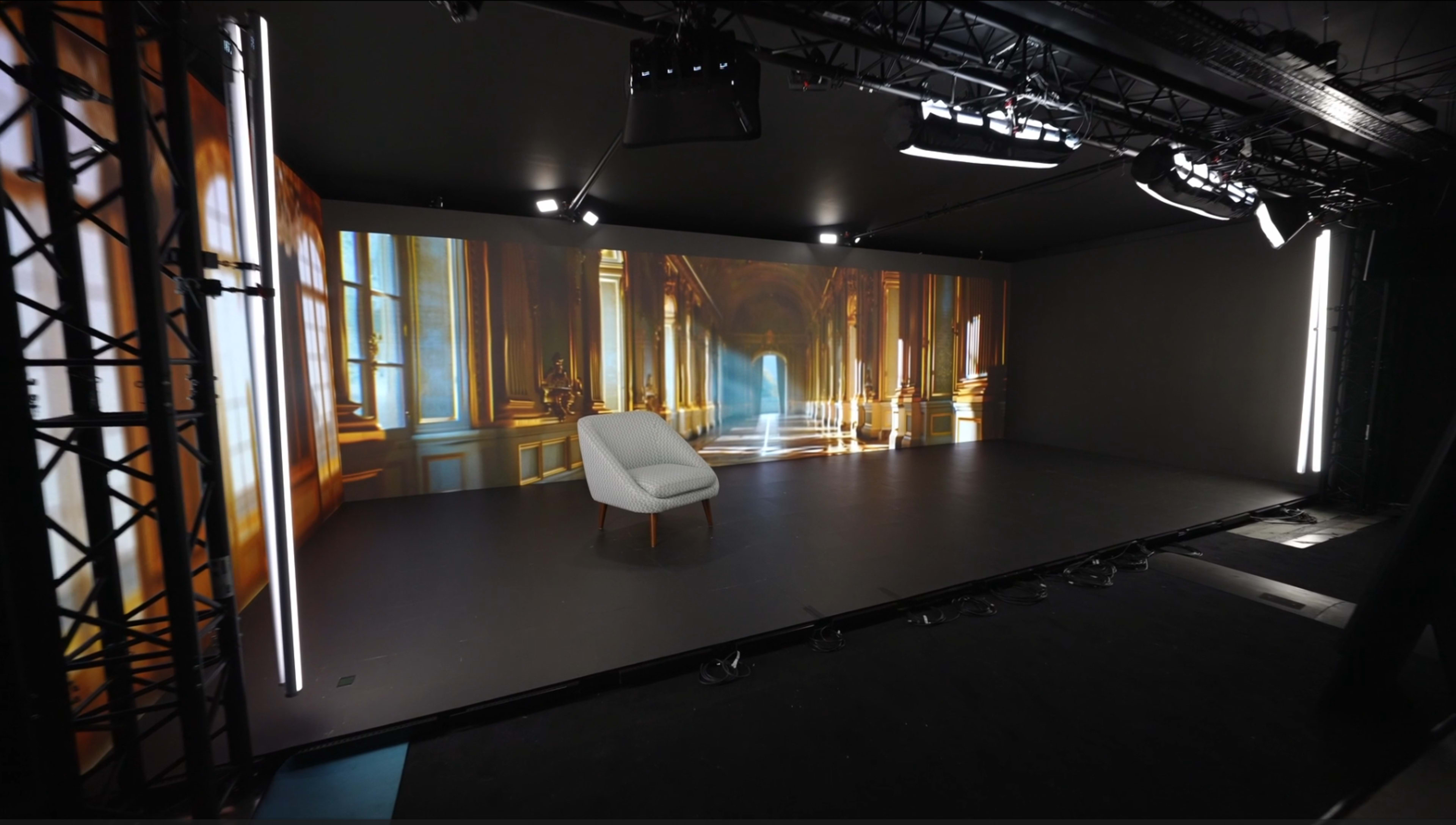 The image shows a modern studio space featuring a single white chair in front of a large backdrop with a digitally rendered interior scene of windows and ornate architecture.