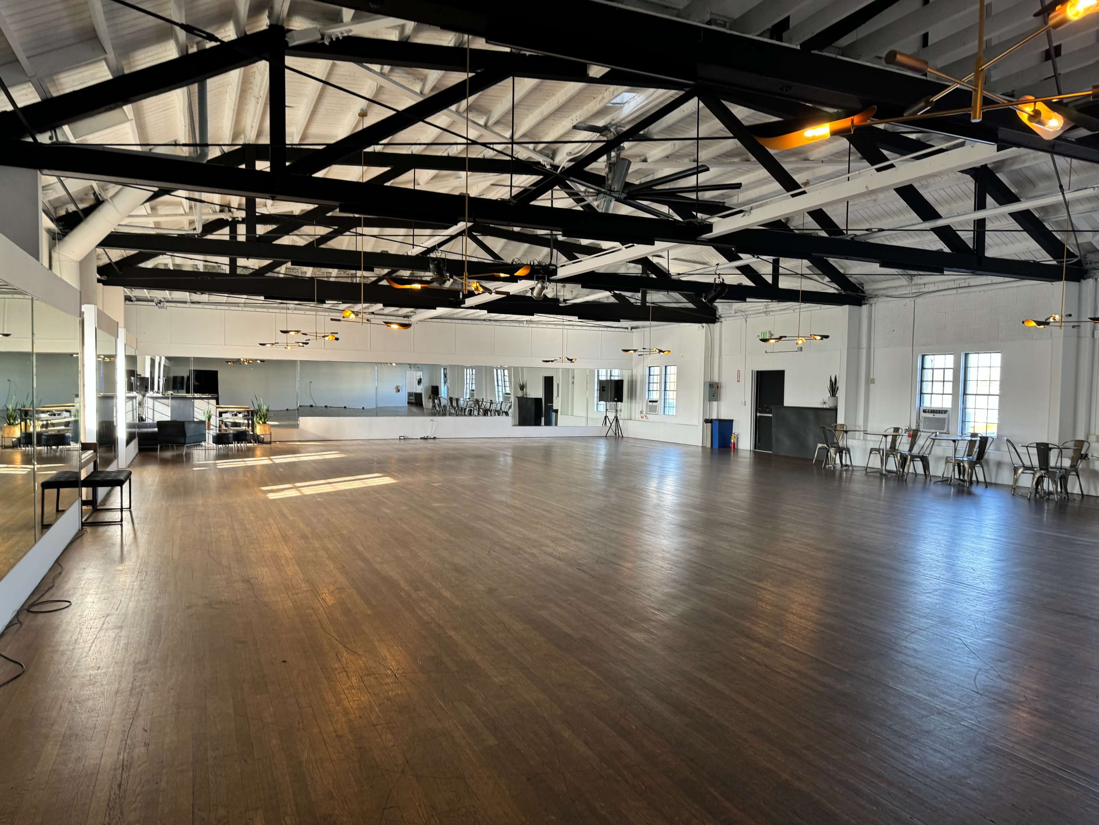 The image shows a spacious, well-lit dance studio with wooden flooring, large mirrors, and several ceiling fans.