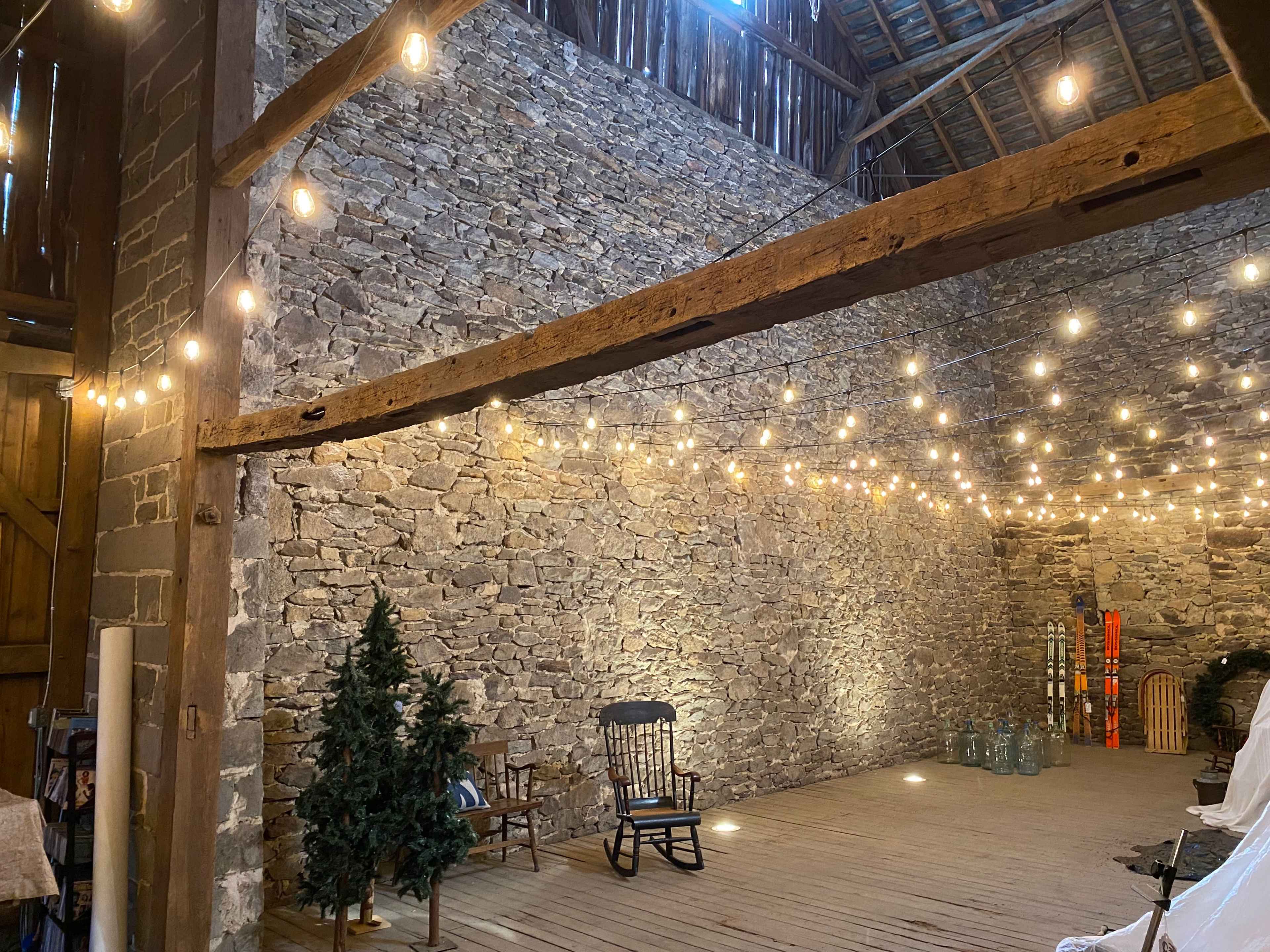 A rustic barn interior with a stone wall, wooden beams, and string lights hanging overhead.