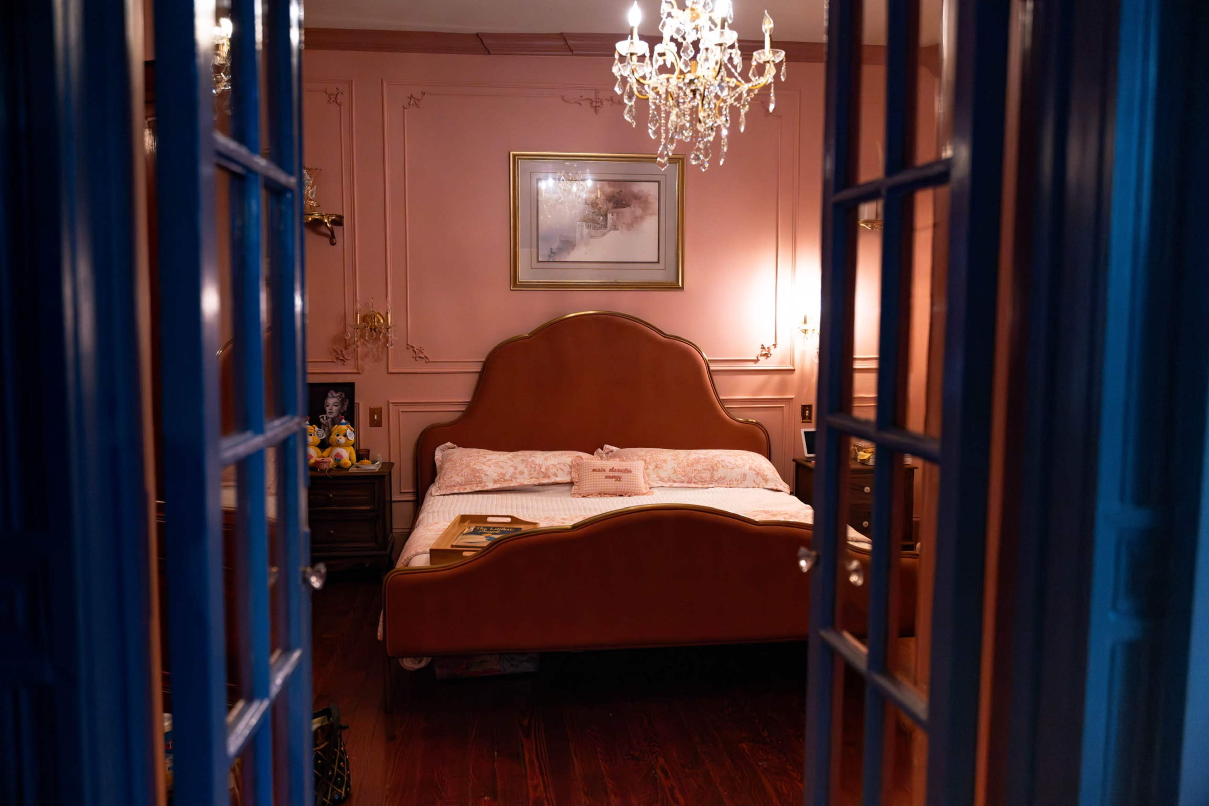 The image shows a view of a bedroom with a large, curved headboard bed, pink walls, and a chandelier.