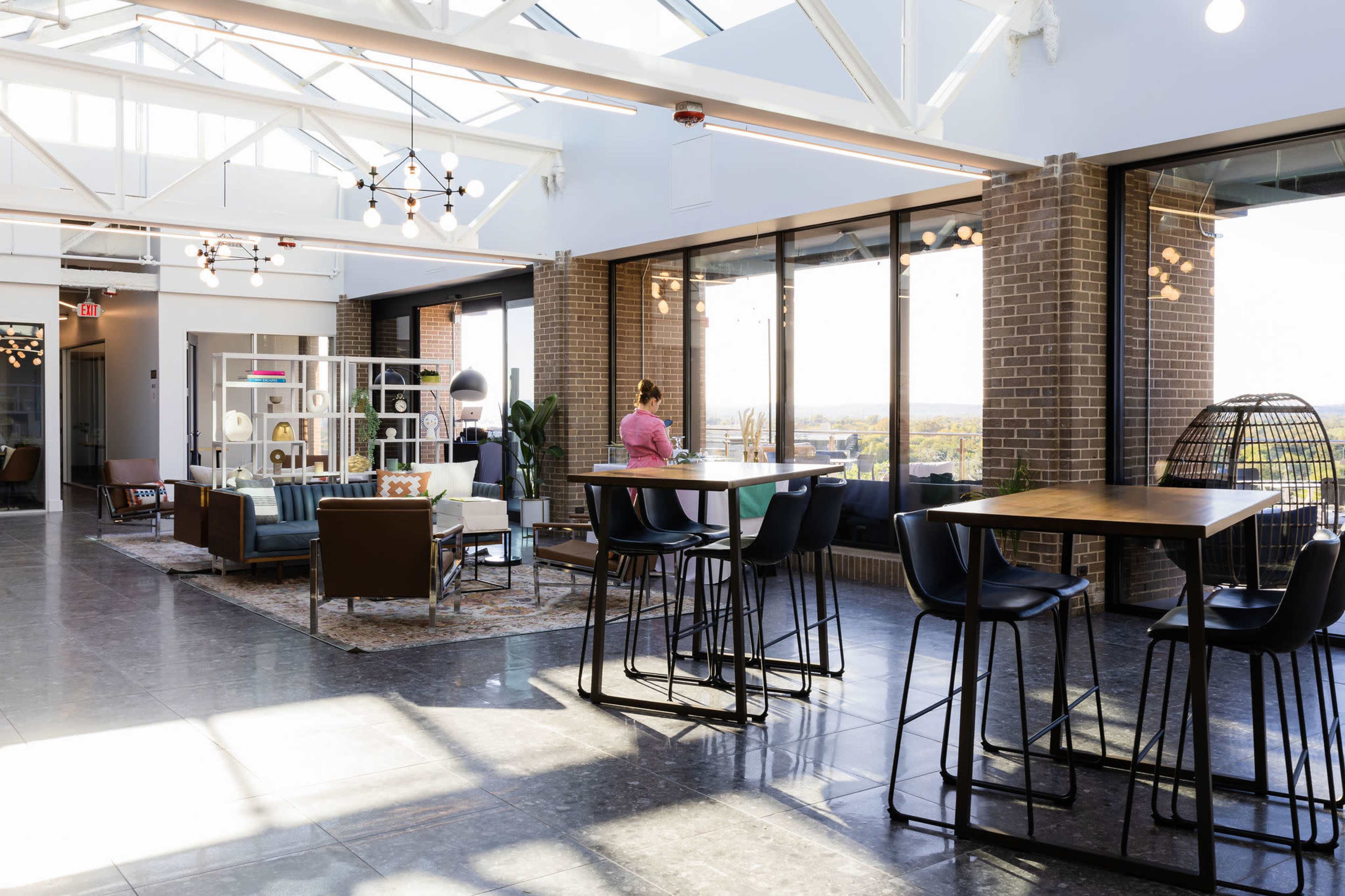 The image shows a modern, open-concept lounge area with large windows, stylish furniture, and a high ceiling.