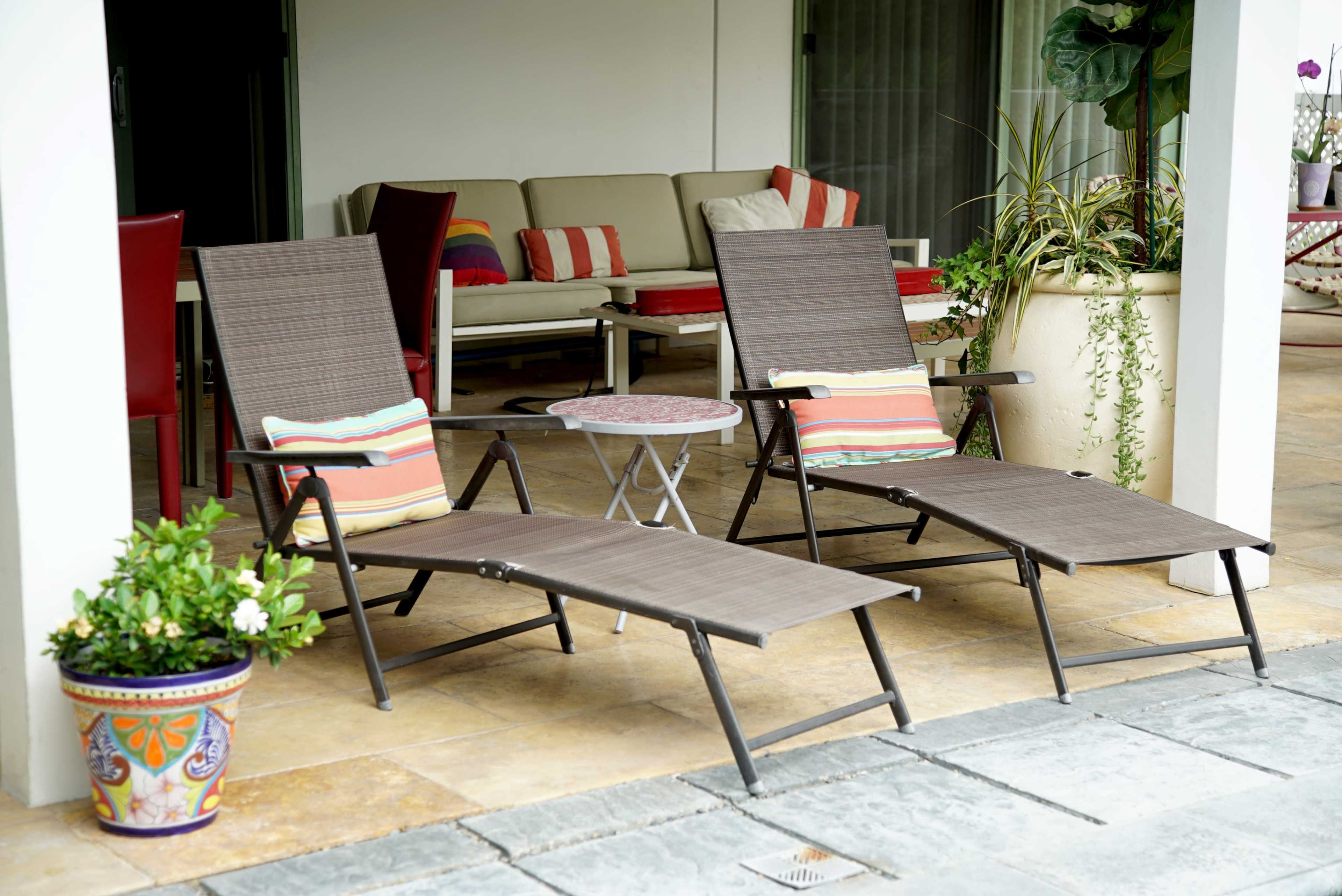 Two reclining lounge chairs with cushions sit beside a small table on a patio covered by a roof, adjacent to a sofa and decorative plants.
