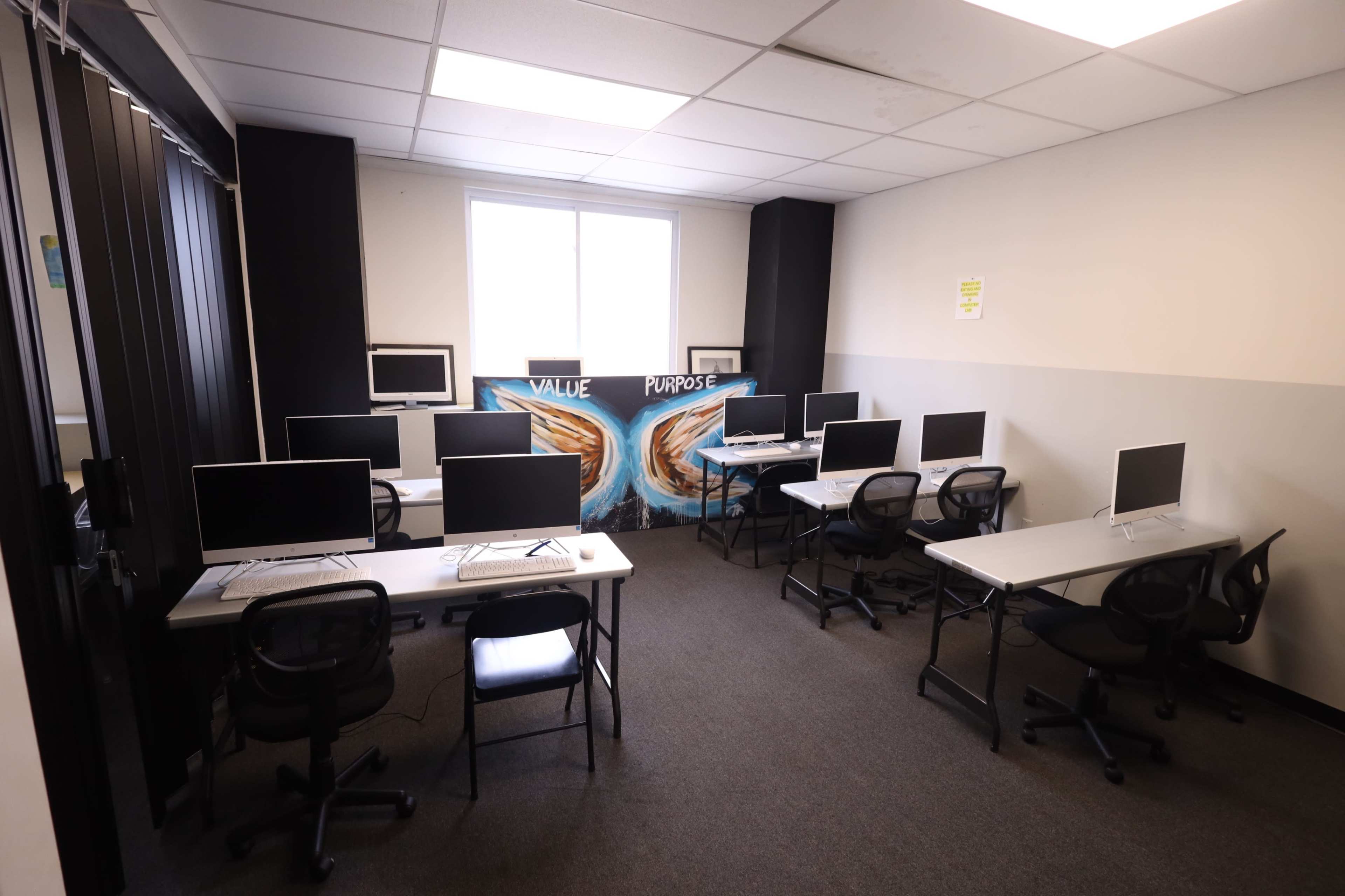 The image shows a classroom equipped with multiple computer desks arranged in a U-shape, with a mural featuring the words "VALUE" and "PURPOSE" on the back wall.