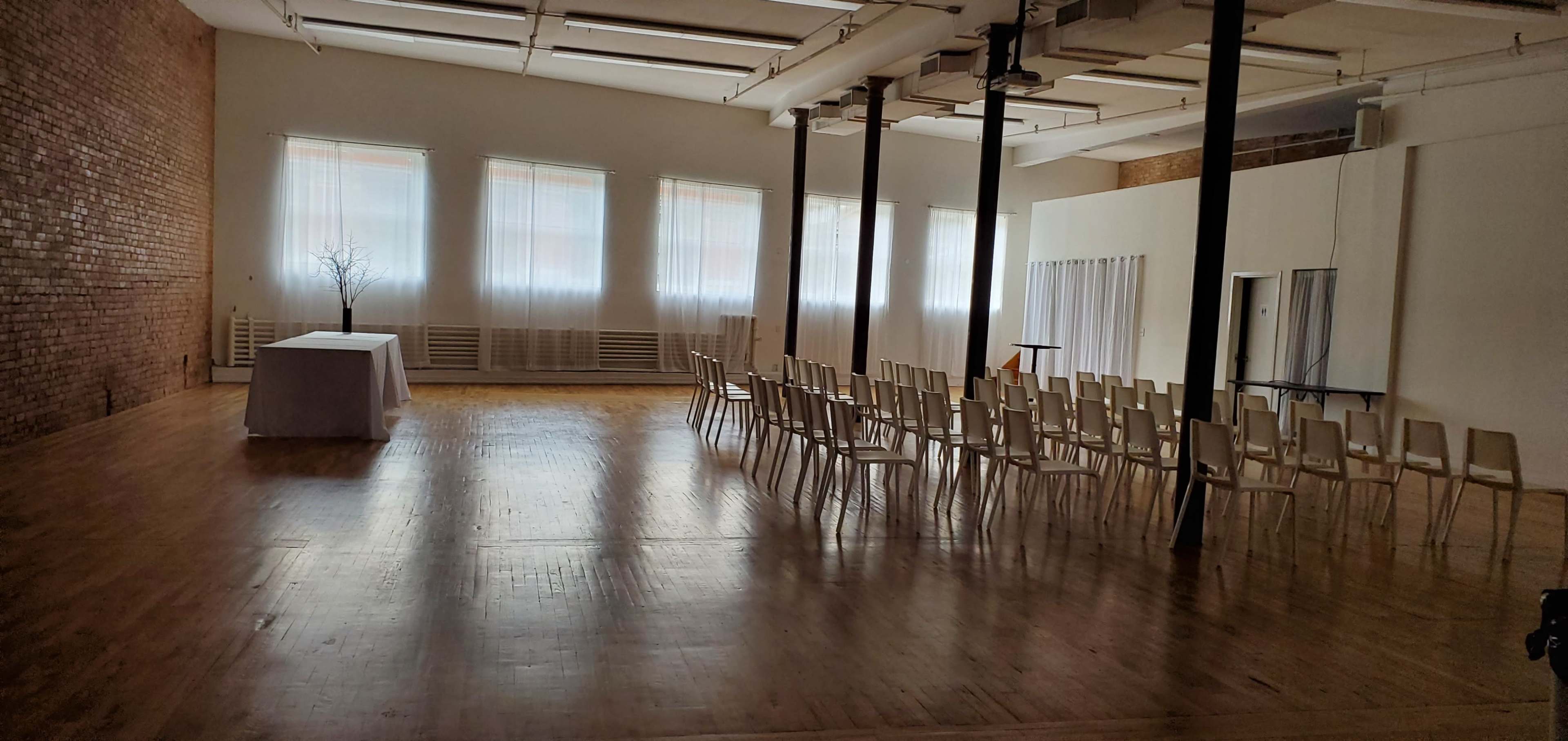 The image shows a spacious, vacant room with wooden floors, white walls, and rows of white chairs facing a simple table at the front.